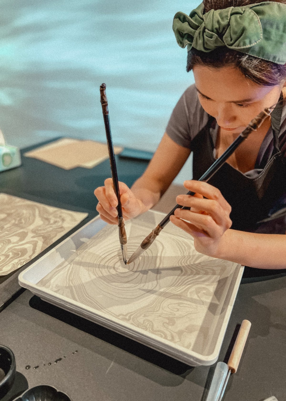 A woman wearing a green scrunchie and dark apron is shown from the side, actively painting a marbled pattern in a square ceramic tray using two paintbrushes.