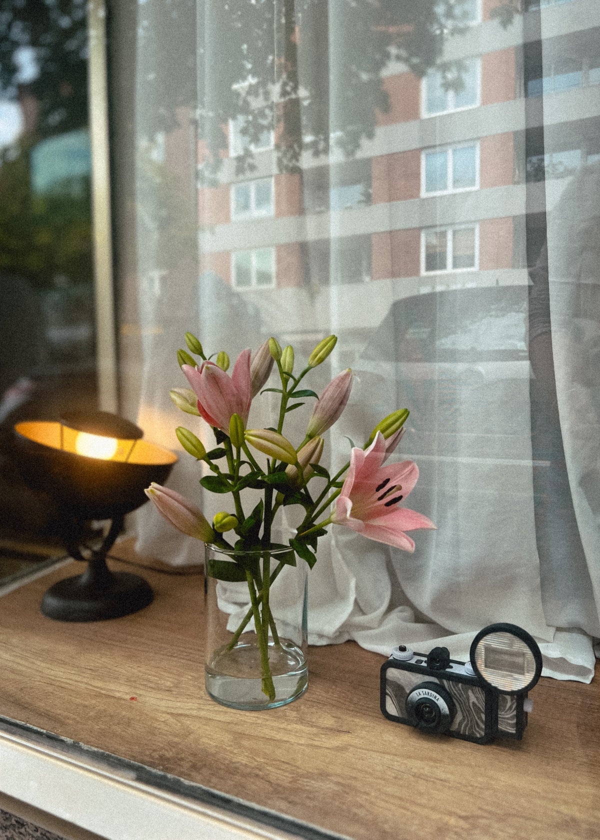 Pink lilies in a clear glass vase on a wooden windowsill, with a lit candle, a vintage camera, and a window view of a building in the background.