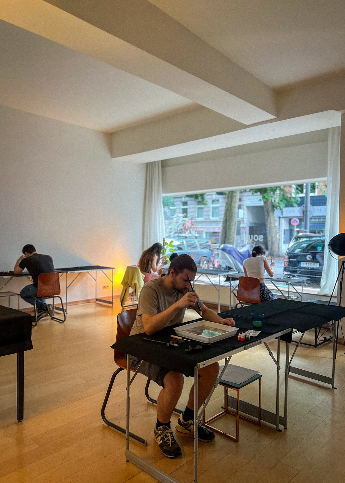A man sits at a black table in a bright, modern cafe or studio space with wood floors and large windows. Other people are seated at tables in the background, and a street scene is visible outside the window.