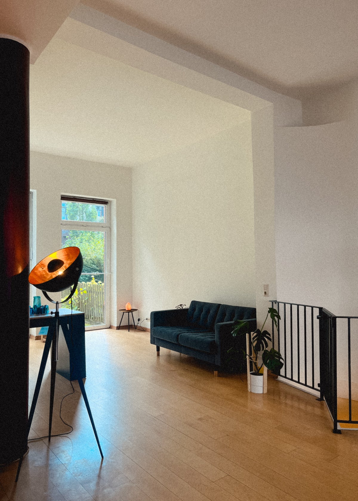 A modern interior space with light wood floors, white walls, a dark teal sofa, a black metal railing, and a large floor lamp in the foreground.