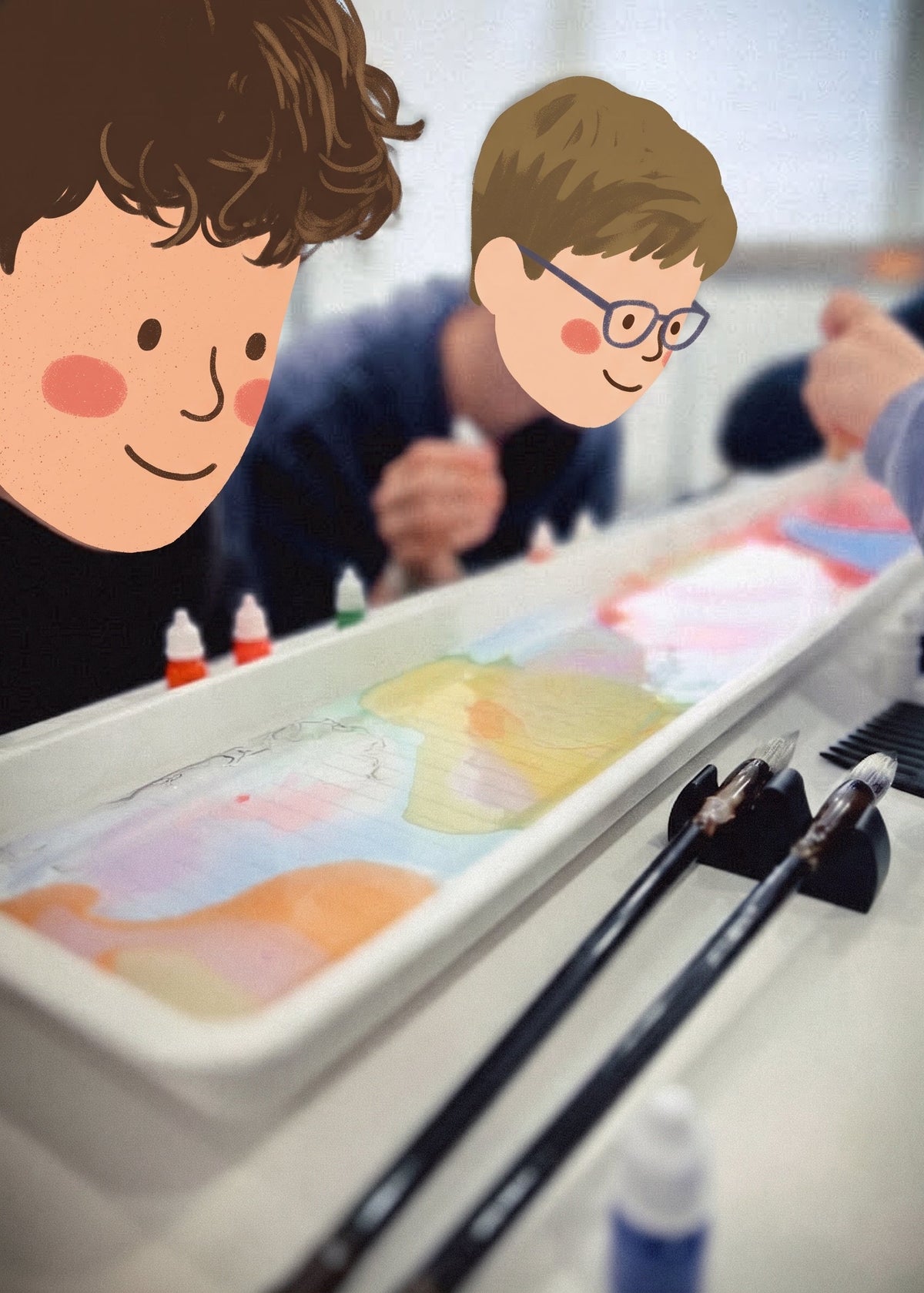 A children's painting or craft activity scene with a long tray holding colorful paint or ink, several paintbrushes, and illustrated cartoon faces overlaid on the left and right sides of the photo.