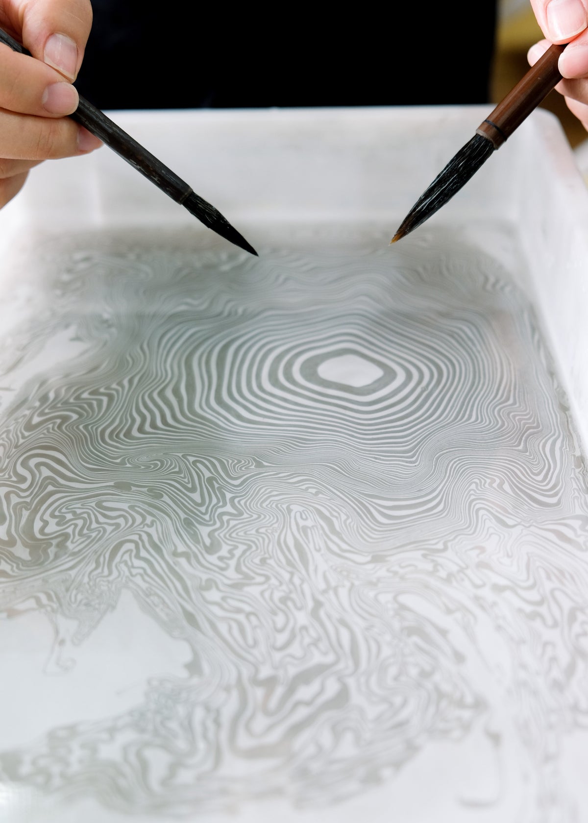 A close-up of a hand-painted topographic map design on a white sheet of paper, with two hands holding traditional paintbrushes hovering over the artwork.