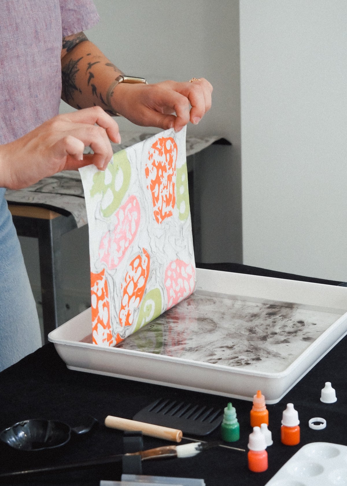 A person holds up a white fabric panel with a colorful abstract pattern of orange, pink, and green shapes, dipping it into a tray of water and ink on a black table. Small bottles of dye or ink and tools are visible on the table.