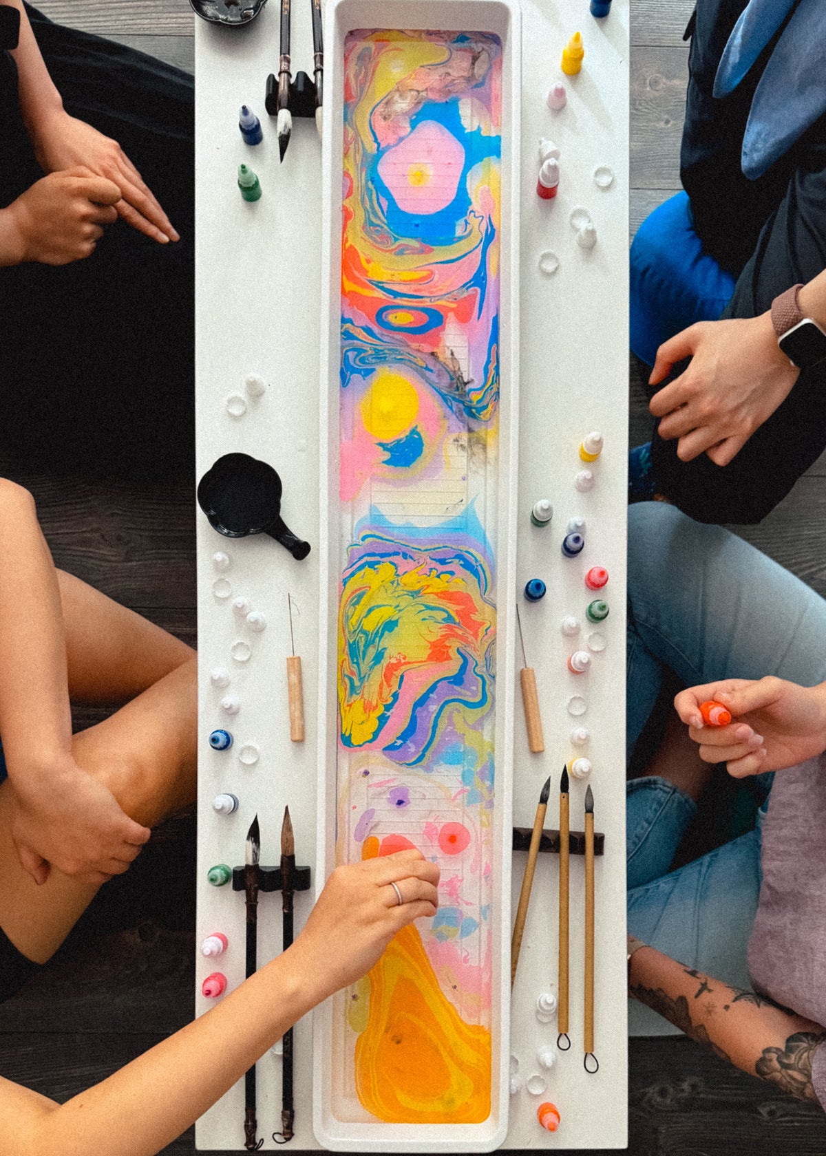 A top-down view of a colorful marbled paint tray surrounded by people's hands and arms as they participate in an art activity. The tray contains vibrant swirls of pink, blue, yellow, and orange paint, with various paint bottles, brushes, and tools arranged around it on a white table.