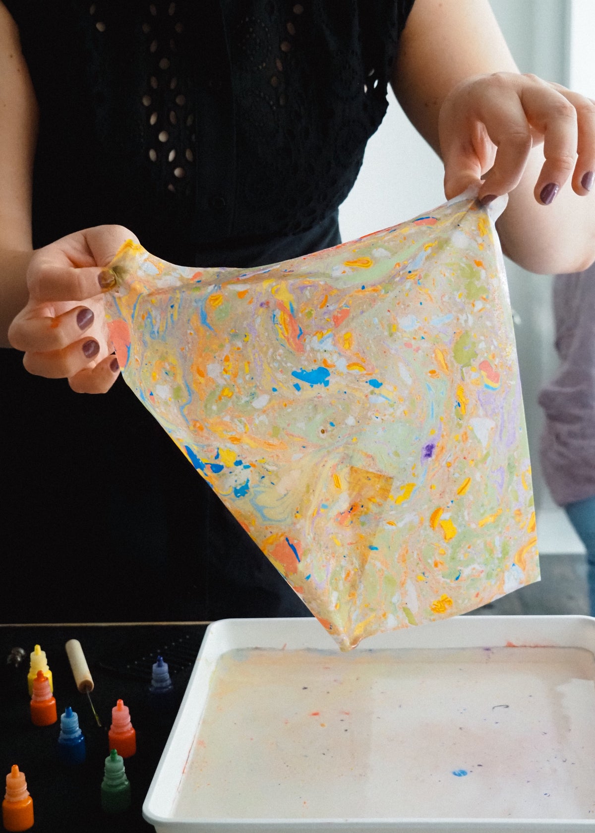 A person holds up a translucent square sheet covered in colorful paint splatters and swirls above a tray of white paint, with small paint bottles on a table below.