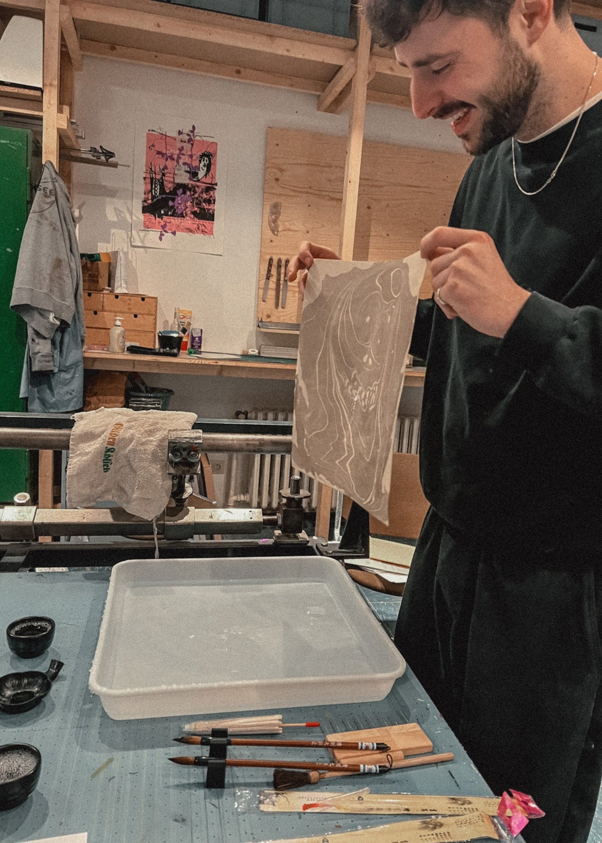 A man in a dark sweatshirt holds up a square piece of white fabric with a wavy pattern, showing it to someone in a creative studio or workshop filled with art supplies, brushes, and wooden panels.