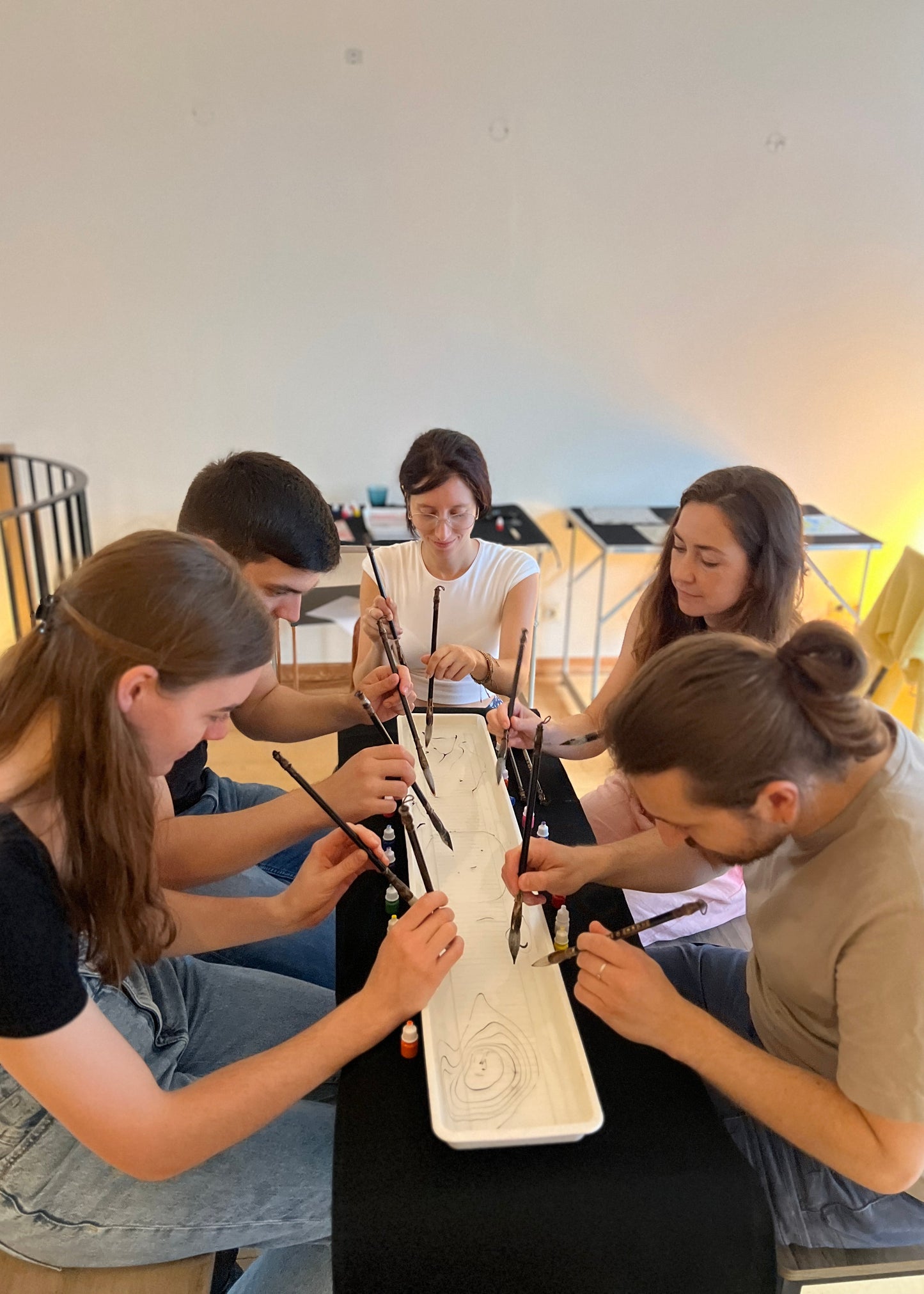 Five people gathered around a table, using calligraphy brushes to carve or write on a white rectangular stone slab in a bright indoor studio or classroom setting.