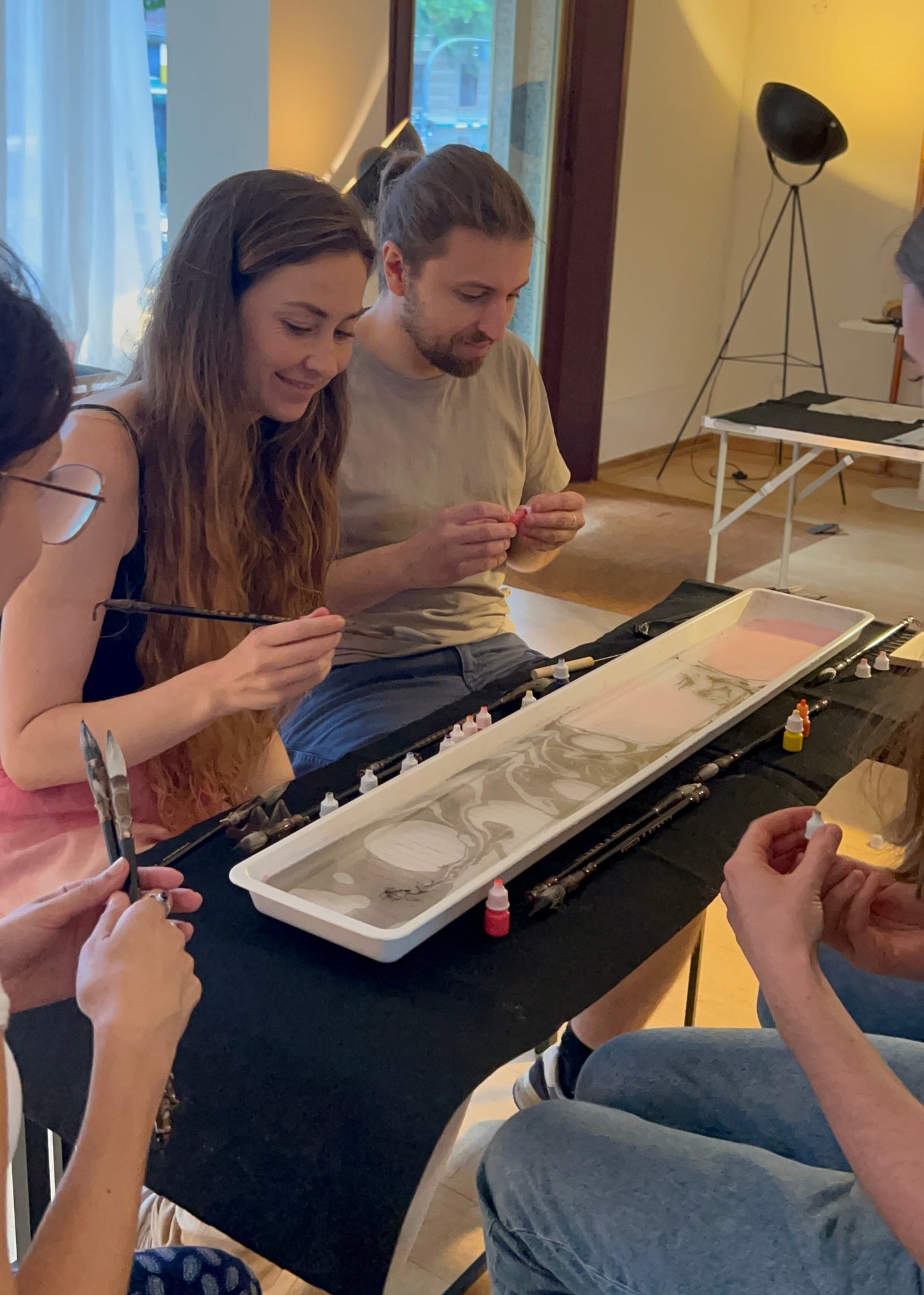 Several people seated around a table in a bright indoor studio space, painting or drawing in a long white rectangular tray with brushes and small paint containers visible on the table.