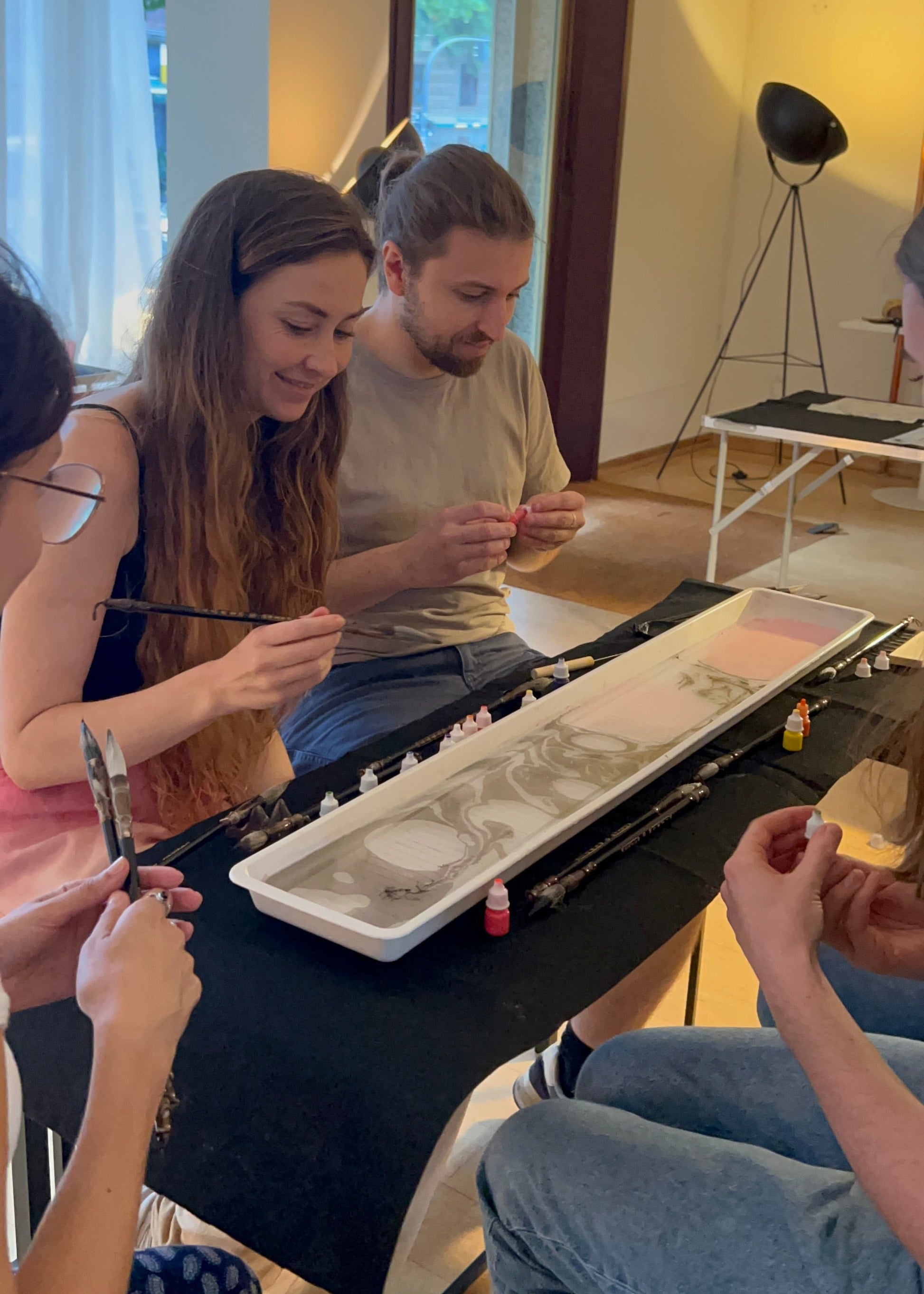 Several people seated around a table in a bright indoor studio space, painting or drawing in a long white rectangular tray with brushes and small paint containers visible on the table.