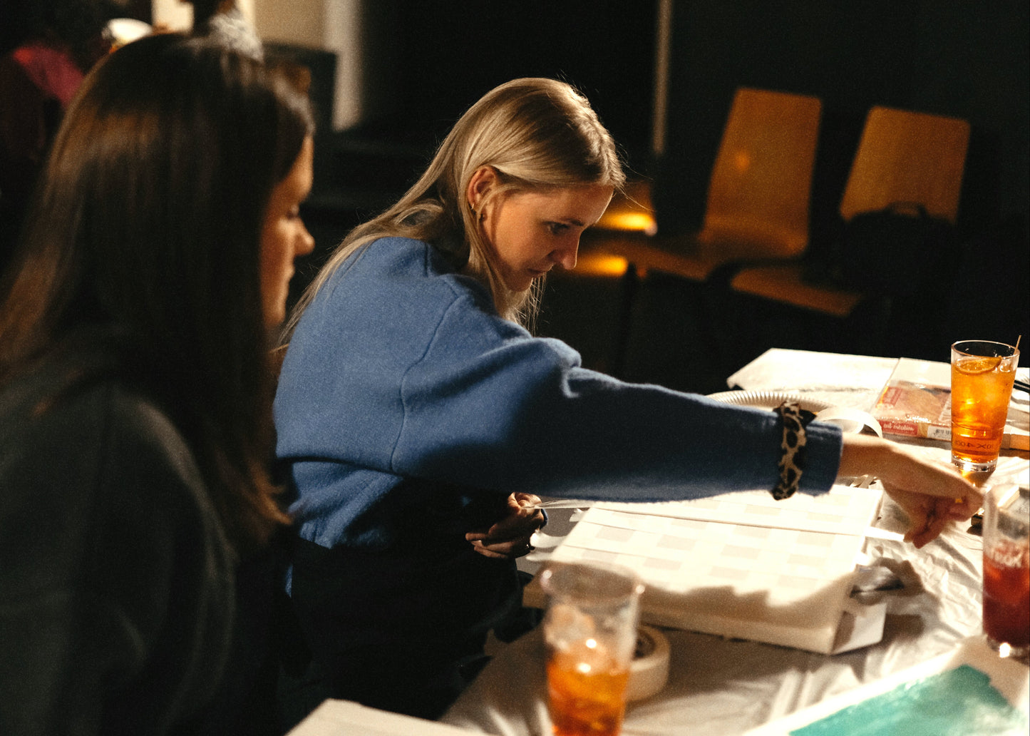 Two women sitting at a table with drinks and papers in a dimly lit room.
