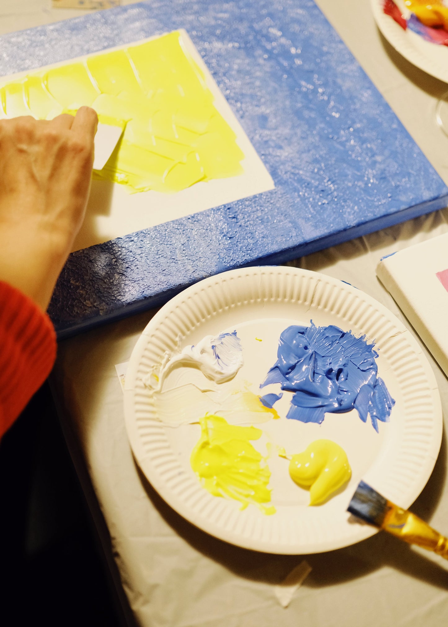 Person painting with yellow and blue on a paper plate, next to a blue paint tray.