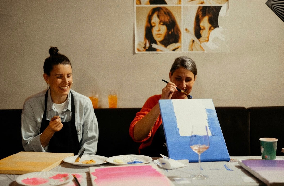 Two women sitting at a table with art supplies, one painting a blue canvas.