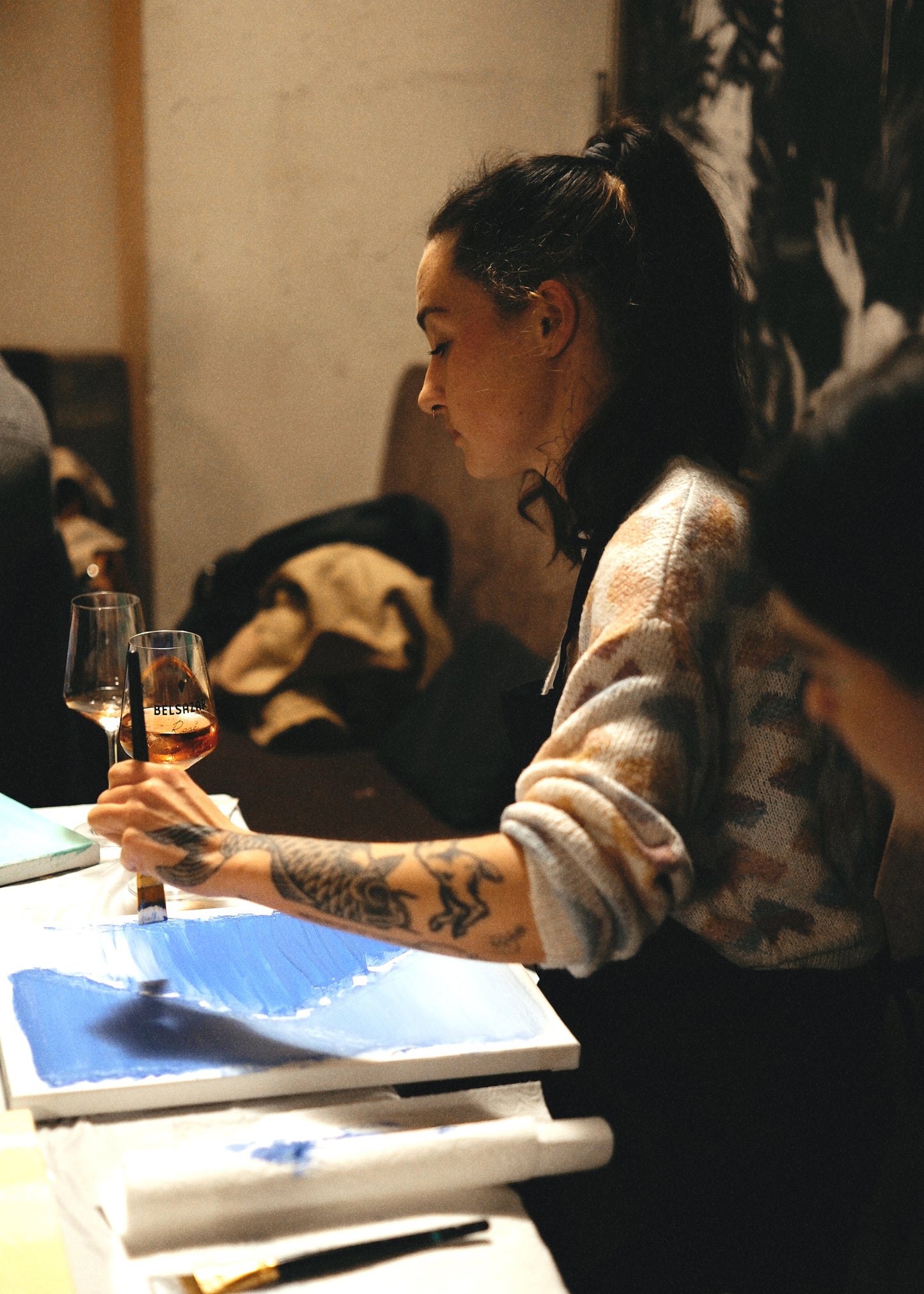 Person with tattoos holding a glass of wine in a dimly lit room