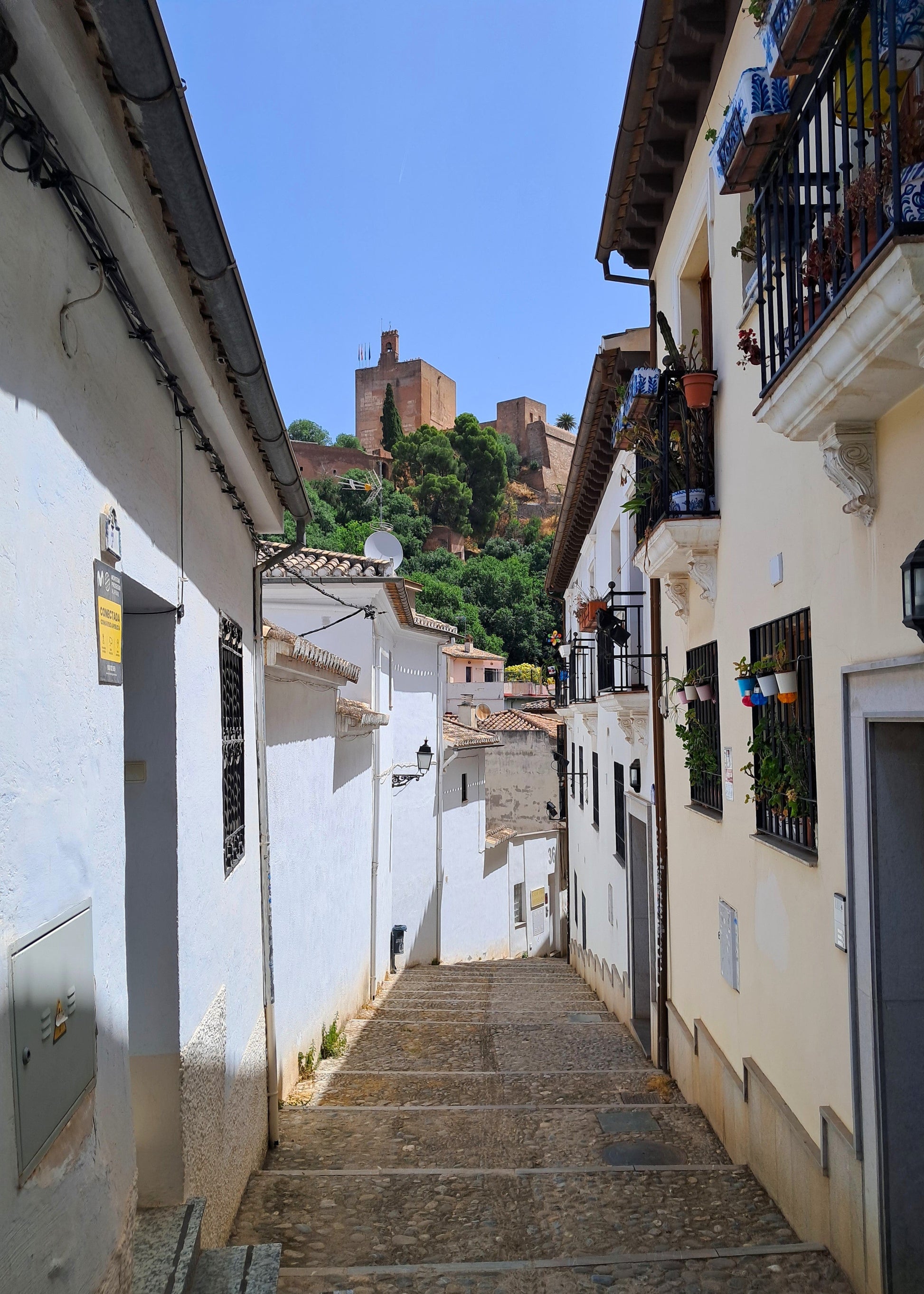 A narrow stone staircase winding between white and beige buildings in a historic town, with terracotta roofs and a castle visible on a green hill under a clear blue sky.