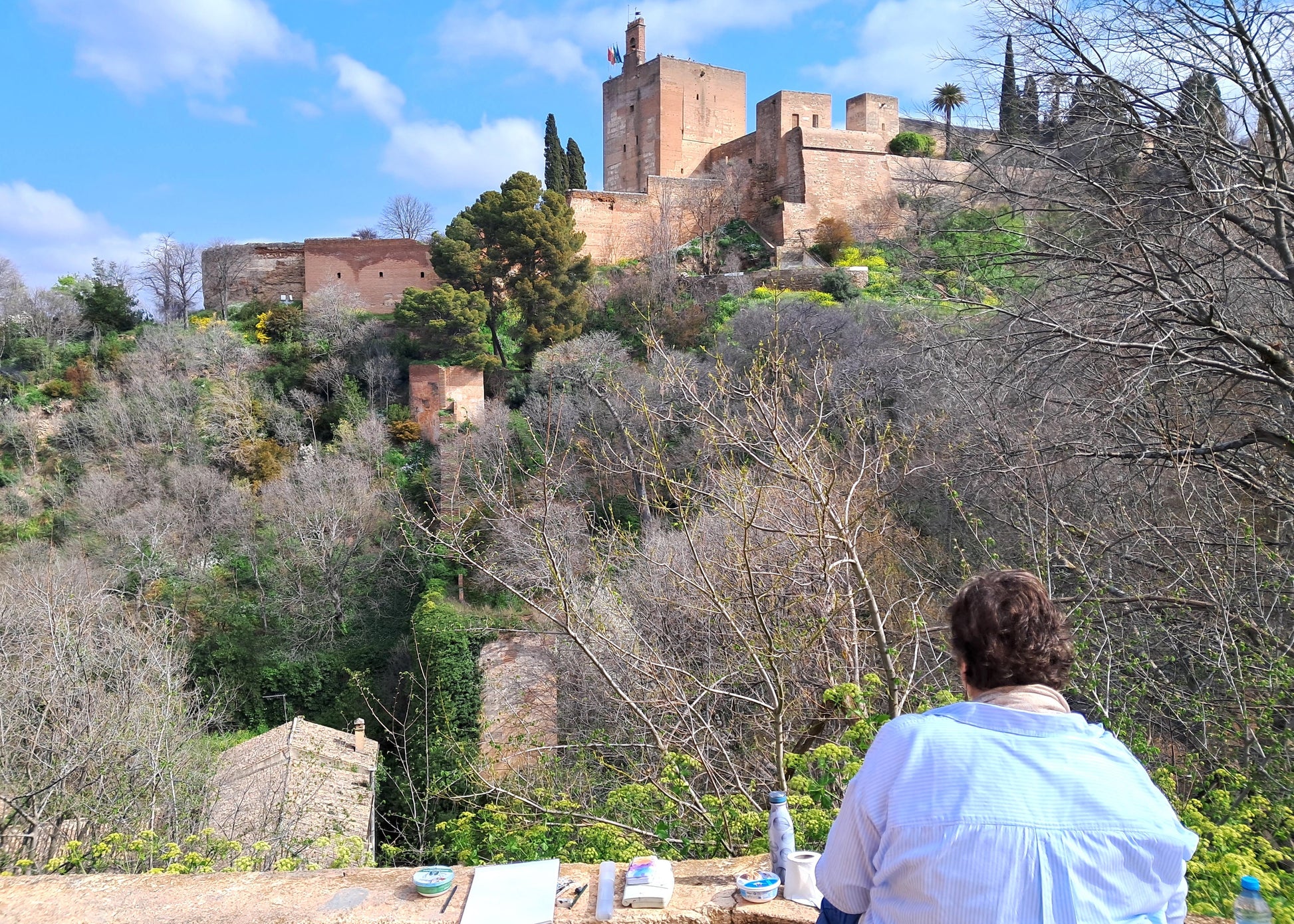 Person painting a scenic view of a castle on a hill with trees and blue sky.