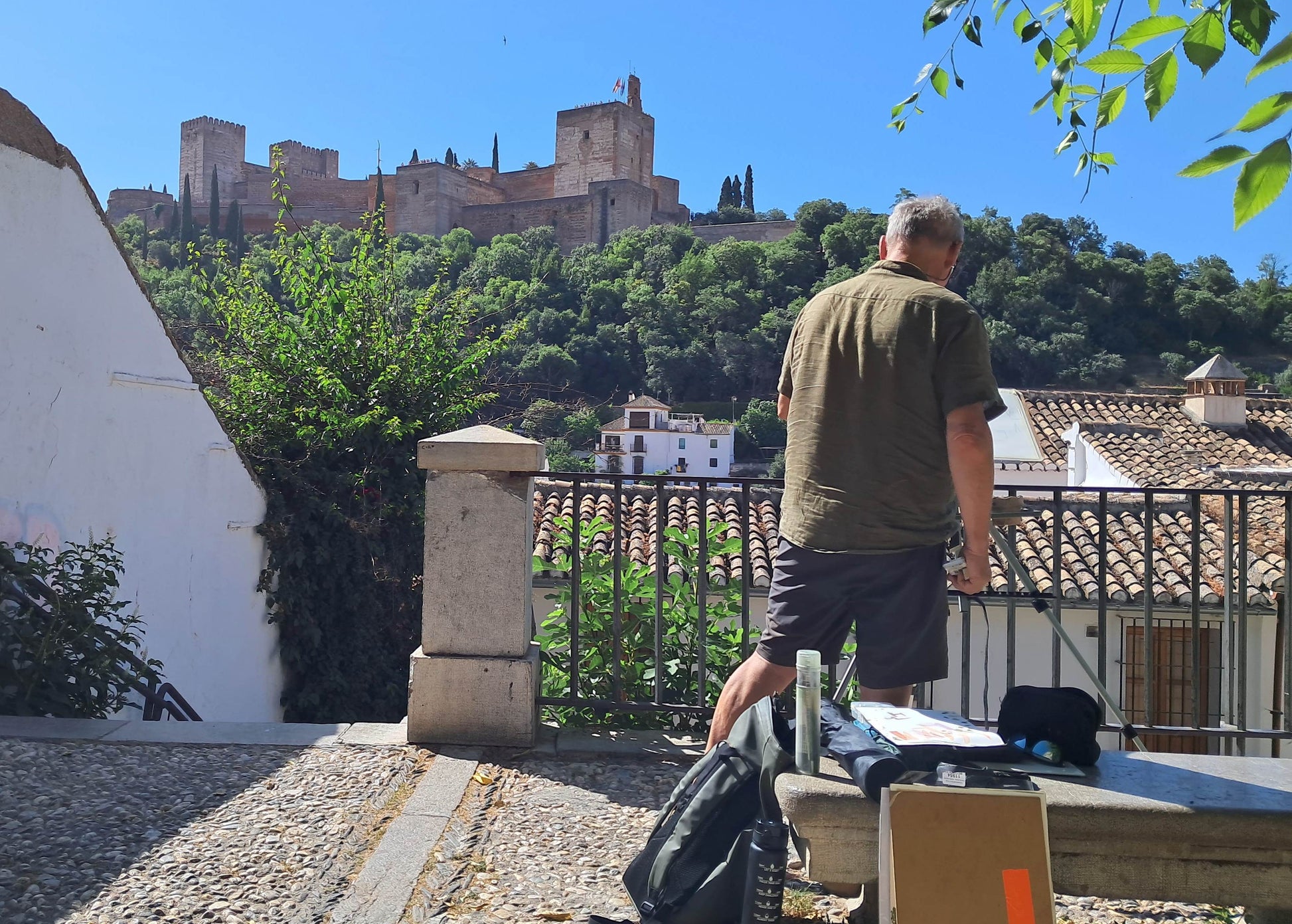 A man stands on a stone bench overlooking a scenic hillside town with a castle on a green hill under a clear blue sky. A cardboard box, a black bag, and papers are on the bench in the foreground.