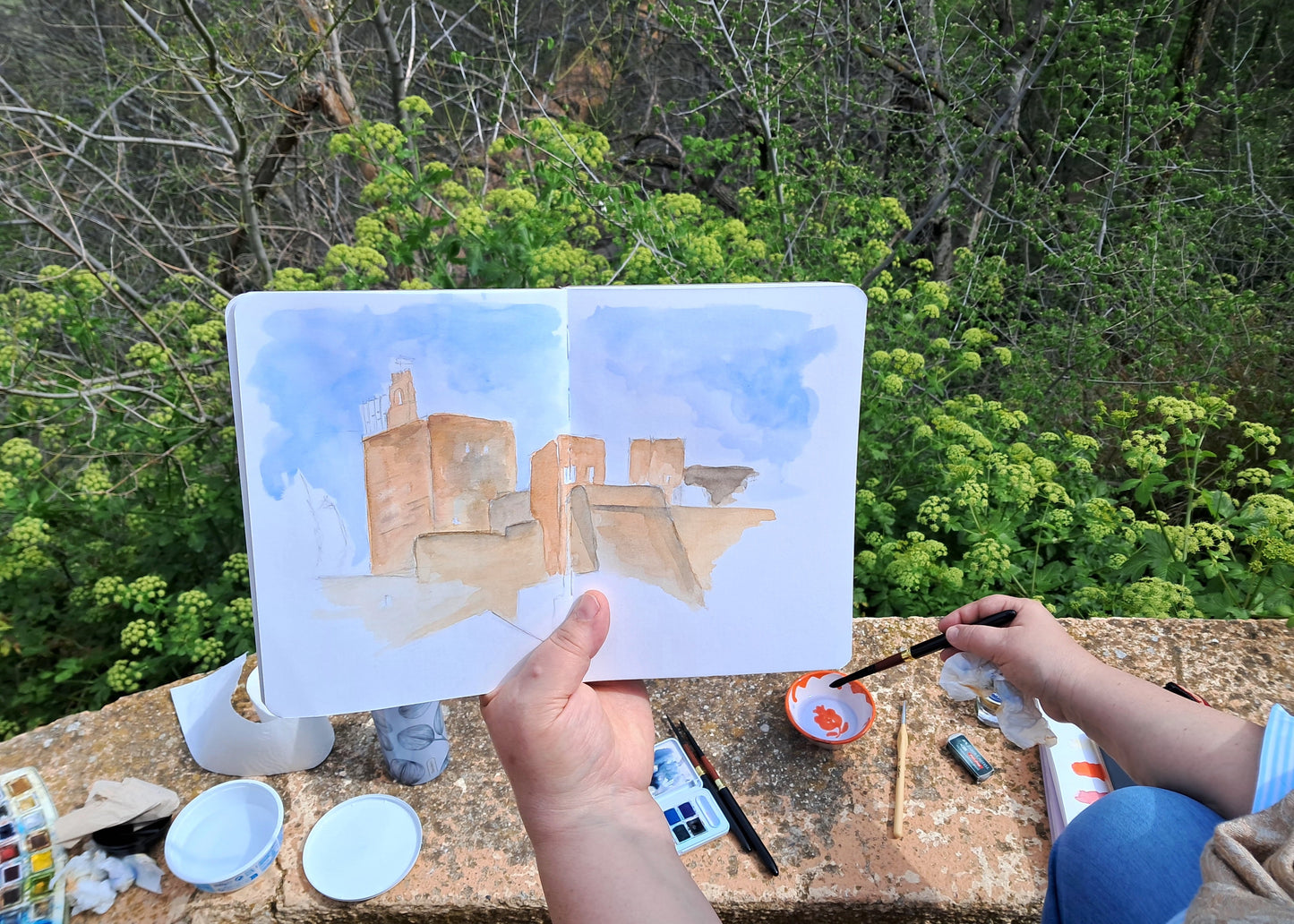 Person painting a cityscape in a sketchbook with natural scenery in the background