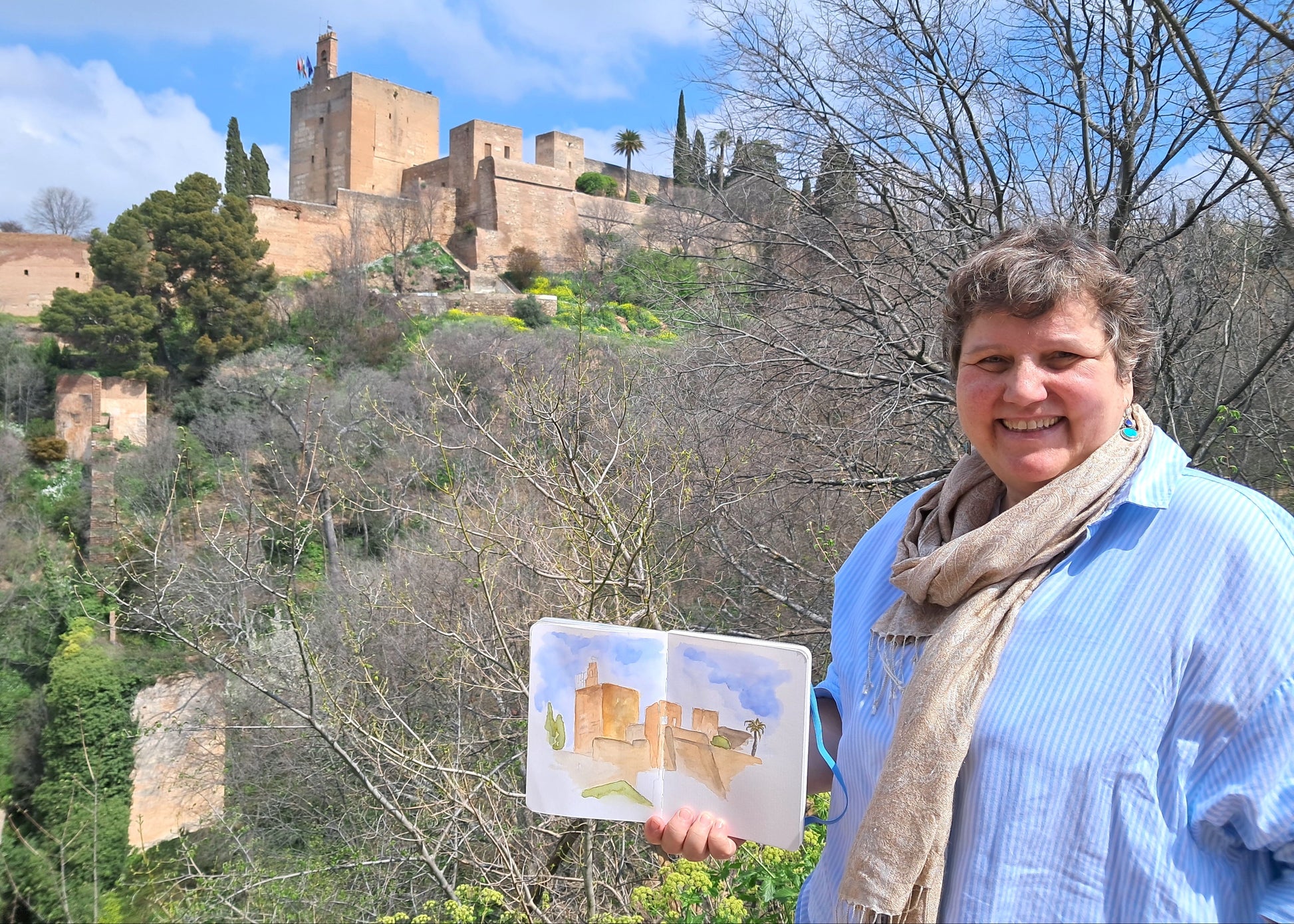 Woman holding a watercolor painting of a castle in front of the actual castle.
