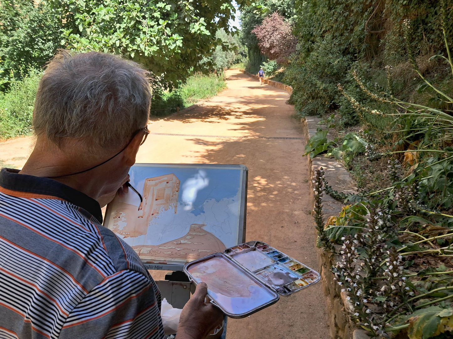 A man with gray hair and glasses, wearing a striped polo shirt, is seen from behind holding an easel with a painted landscape and a small paint palette in a garden pathway setting.