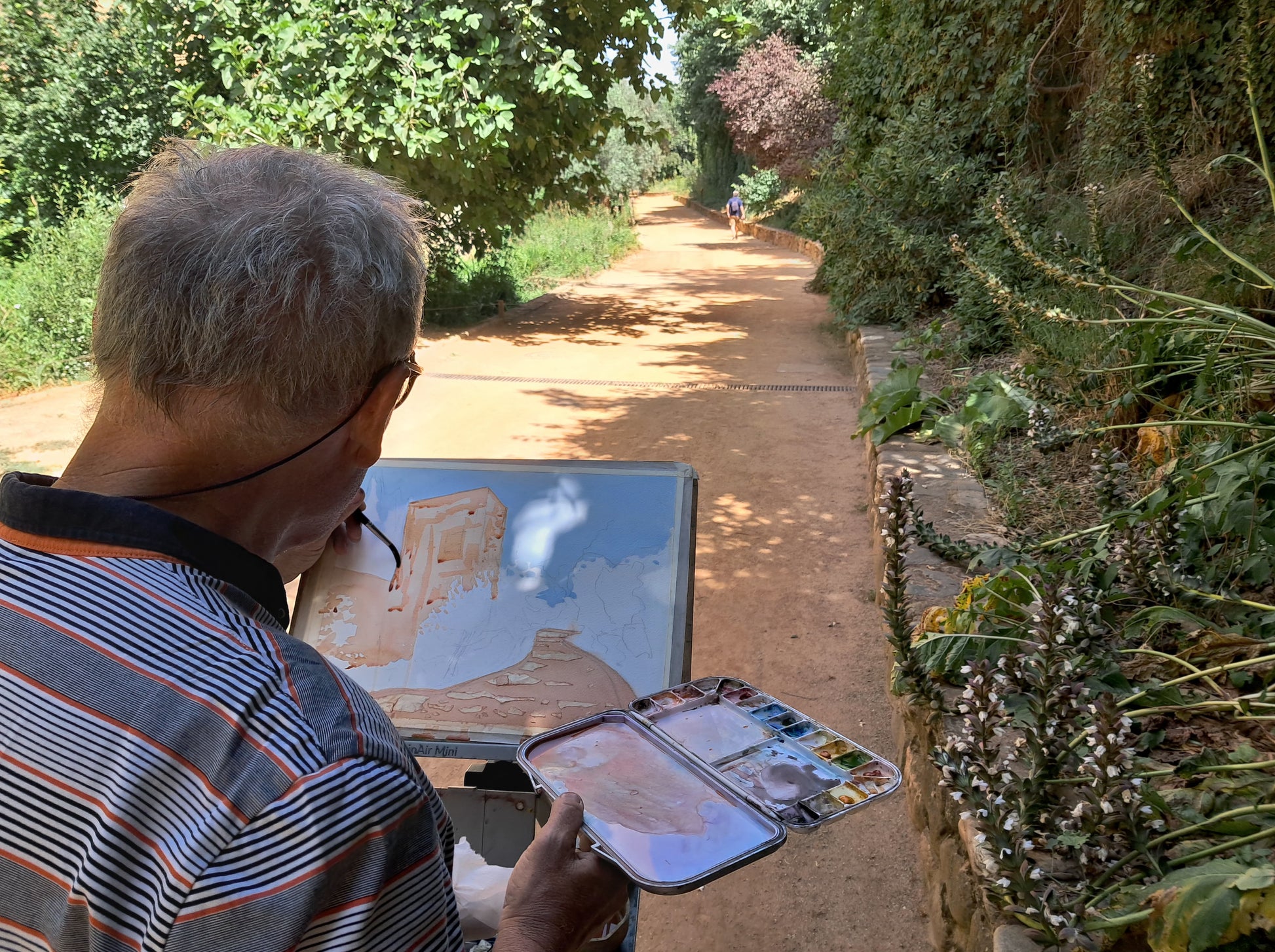 A man with gray hair and glasses, wearing a striped polo shirt, is seen from behind holding an easel with a painted landscape and a small paint palette in a garden pathway setting.