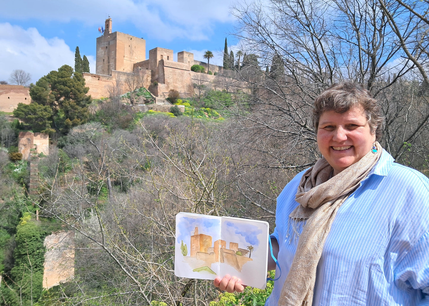 Woman holding a watercolor painting of a castle in front of the actual castle.
