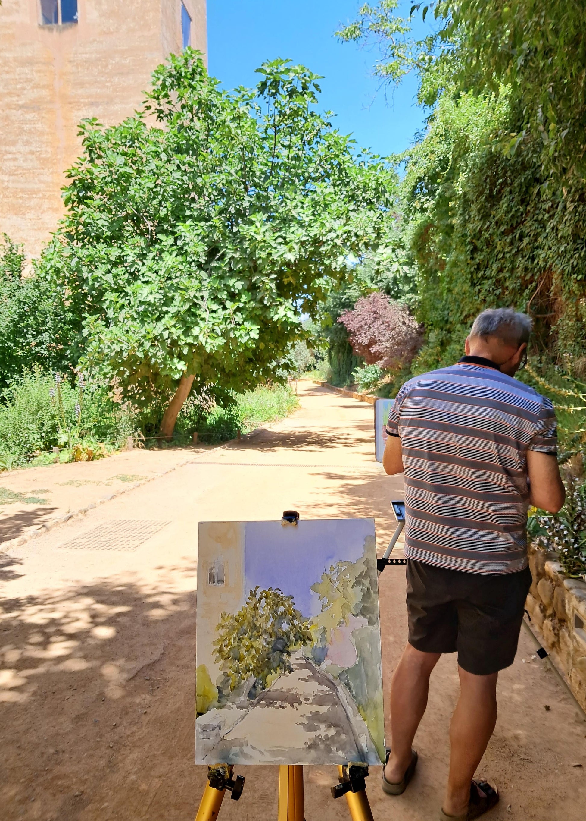 A man stands on a path outdoors, painting a landscape scene on an easel. The scene includes trees, a dirt path, a building wall, and blue sky.