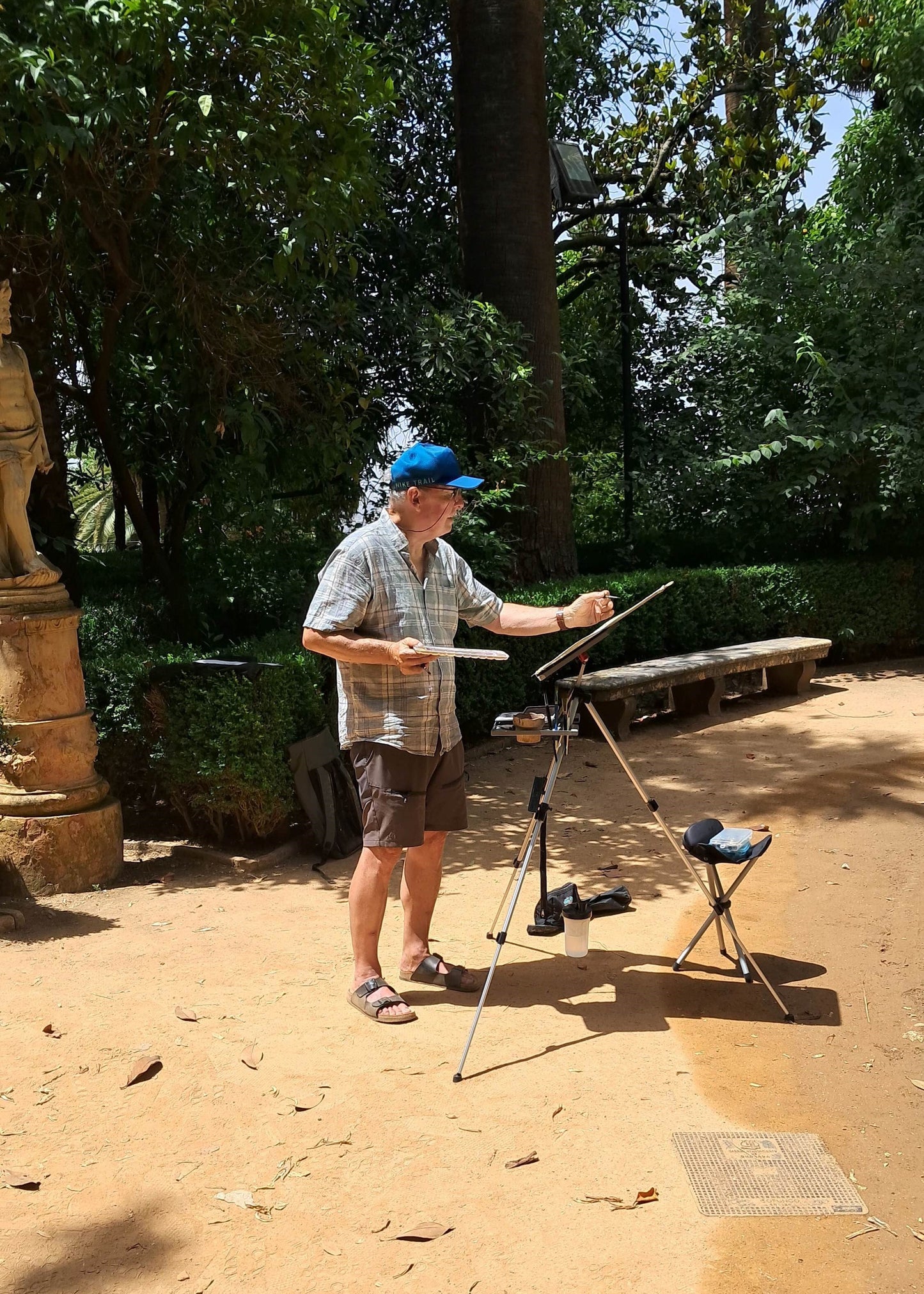 A man stands outdoors in a park-like setting with trees and sandy ground, holding a paintbrush and a palette on a tripod, with a small stool nearby.