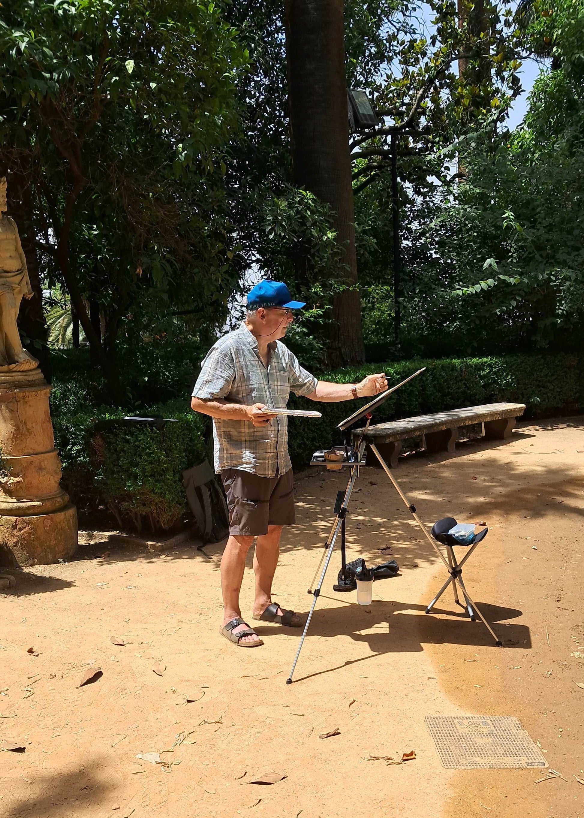 A man stands outdoors in a park-like setting with trees and sandy ground, holding a paintbrush and a palette on a tripod, with a small stool nearby.