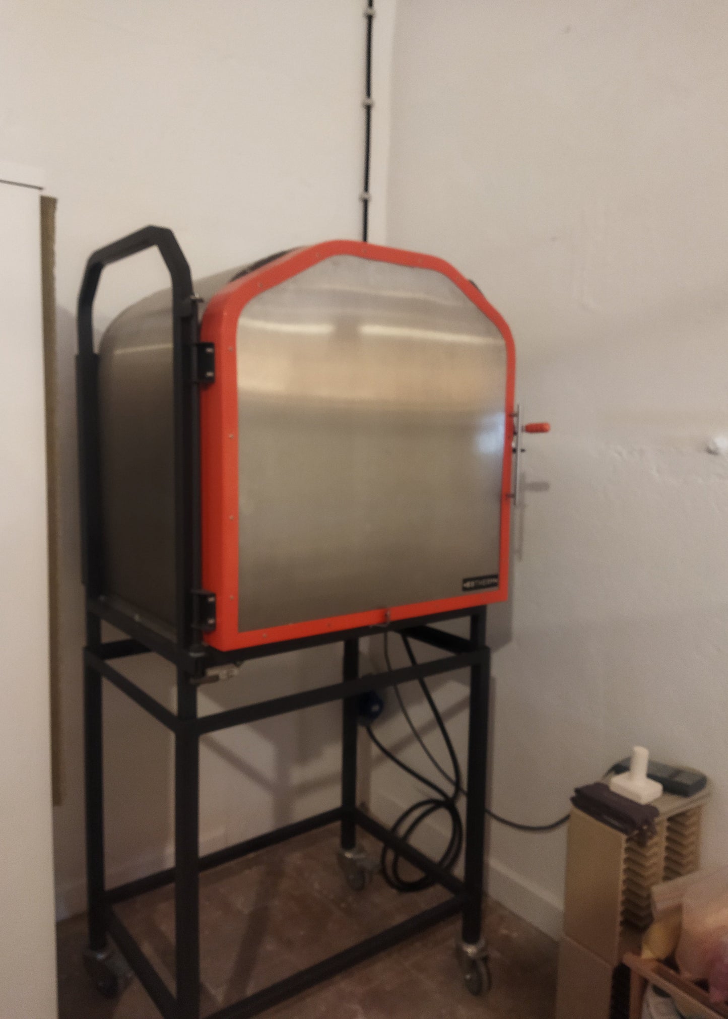 Large metal machine with an orange border on a stand against a white wall.