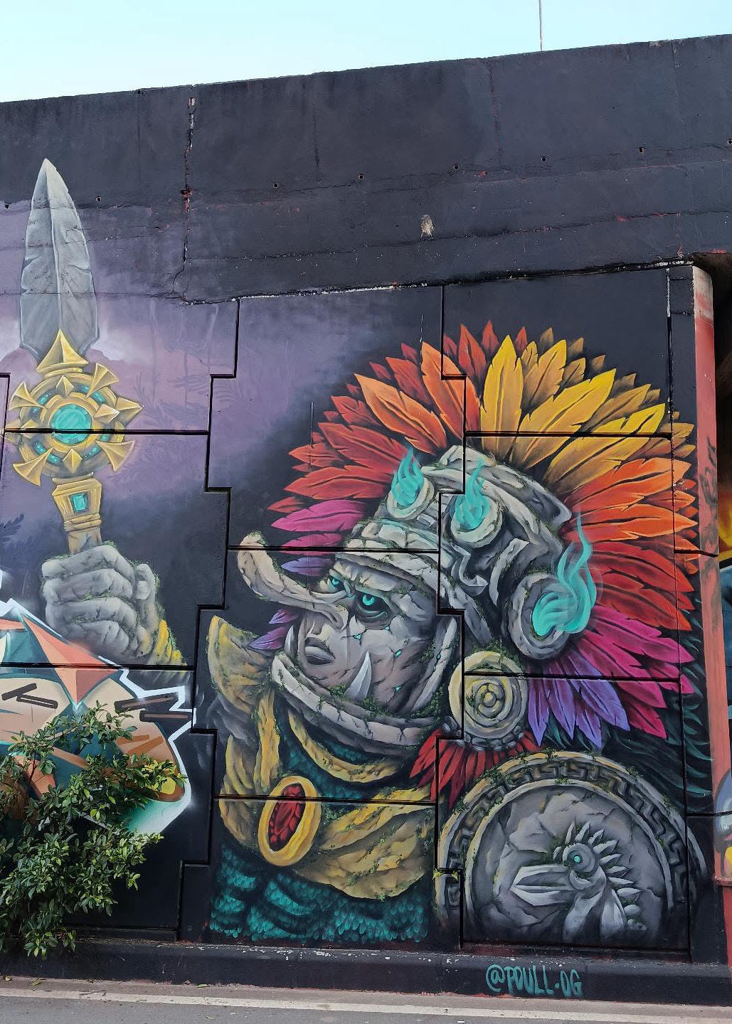 Colorful street art mural painted on a dark wall, featuring a stylized warrior with a bright rainbow feather headdress, gray armor, and a spear, with a small plant growing in a planter at the bottom left.