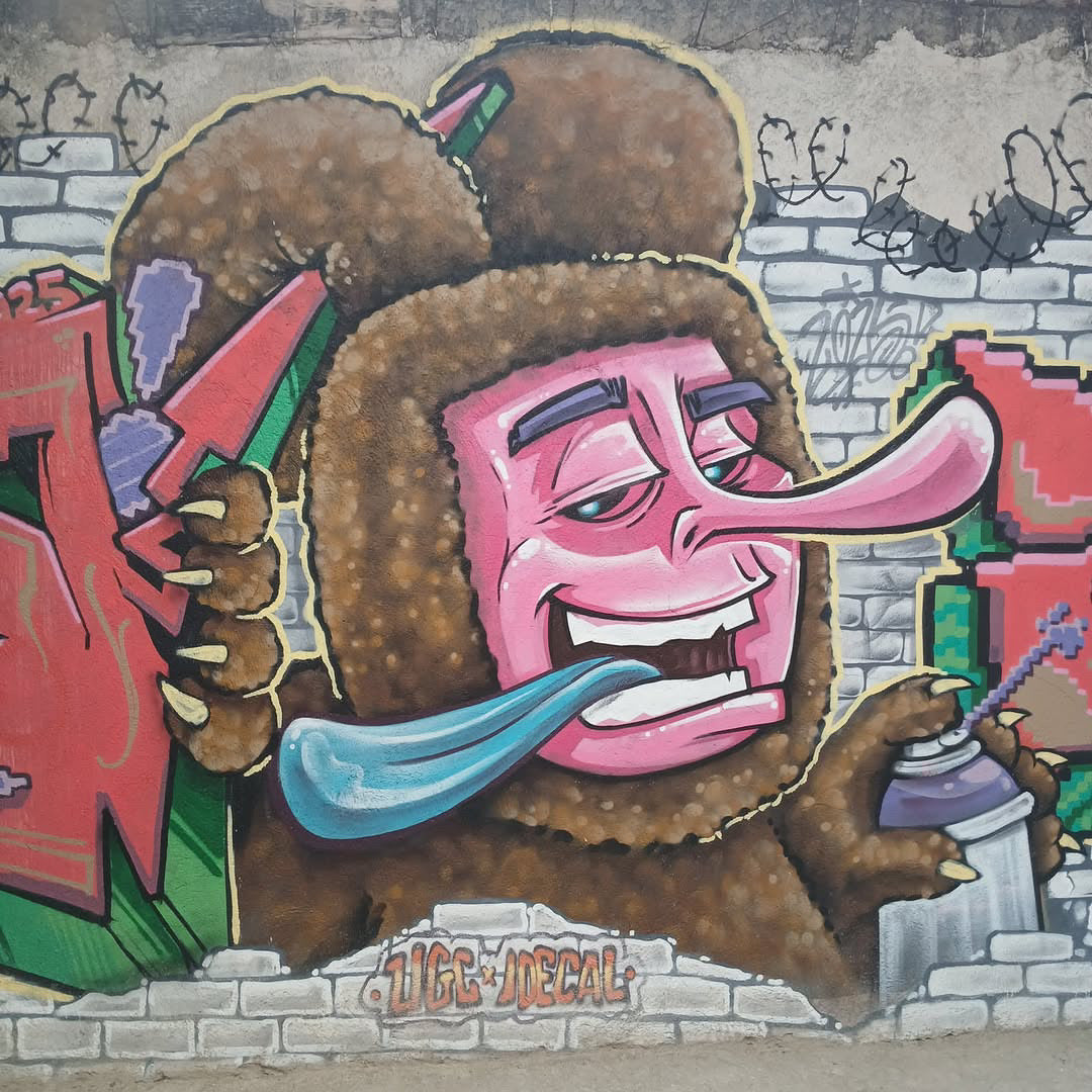 A colorful graffiti-style mural featuring a cartoon character with a brown textured body, large pink face, long pink tongue, and blue tongue, holding spray paint cans against a brick wall background.
