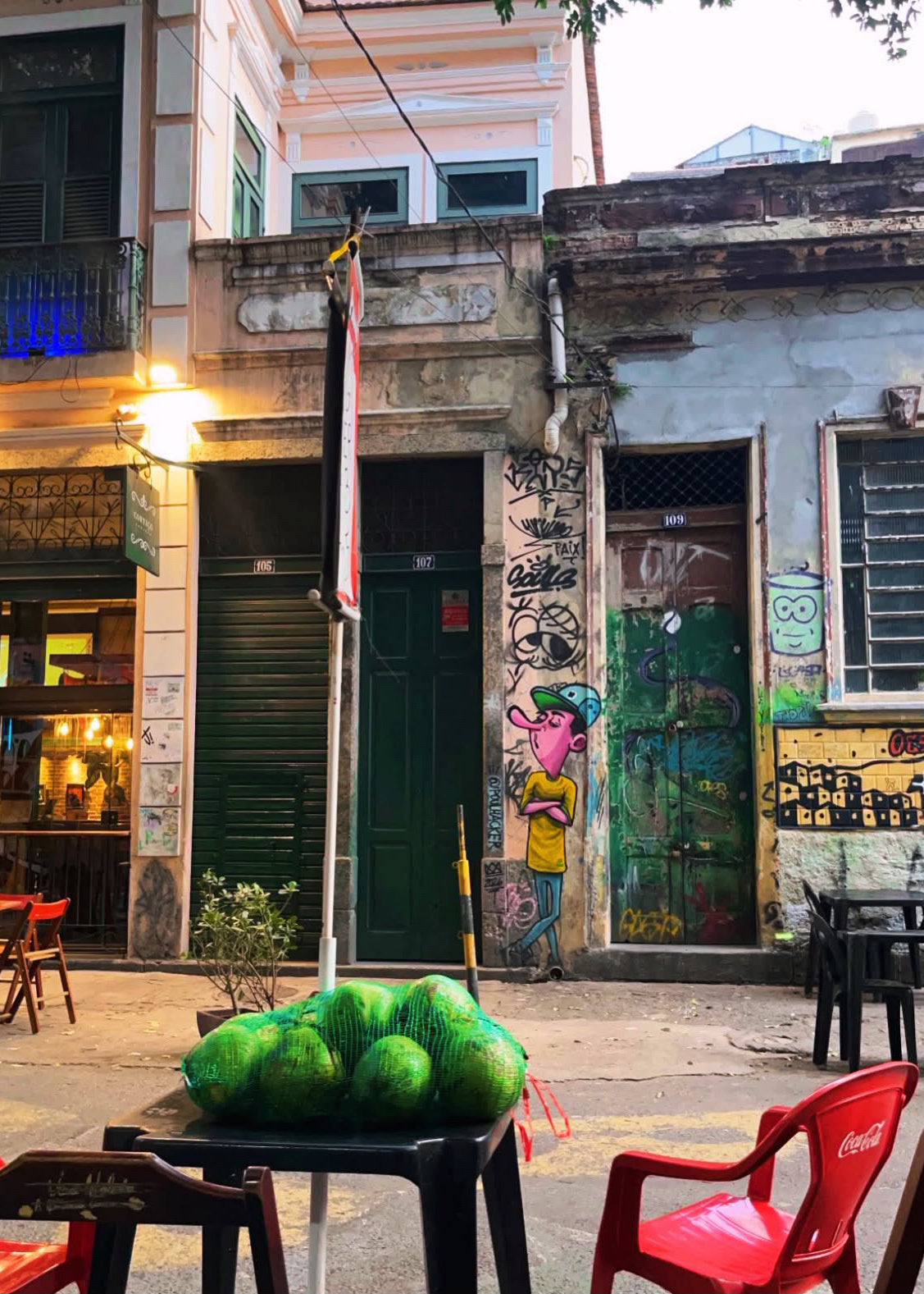 A street scene in an urban alley with old buildings, graffiti-covered walls, outdoor seating, and a table in the foreground holding several green melons.