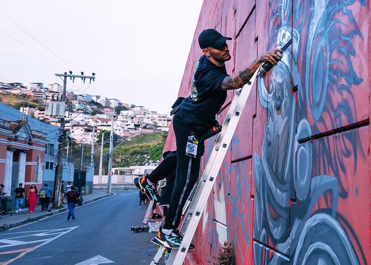 A man in a black cap and black outfit climbs a metal ladder to paint a large red graffiti wall mural in an outdoor urban street setting.
