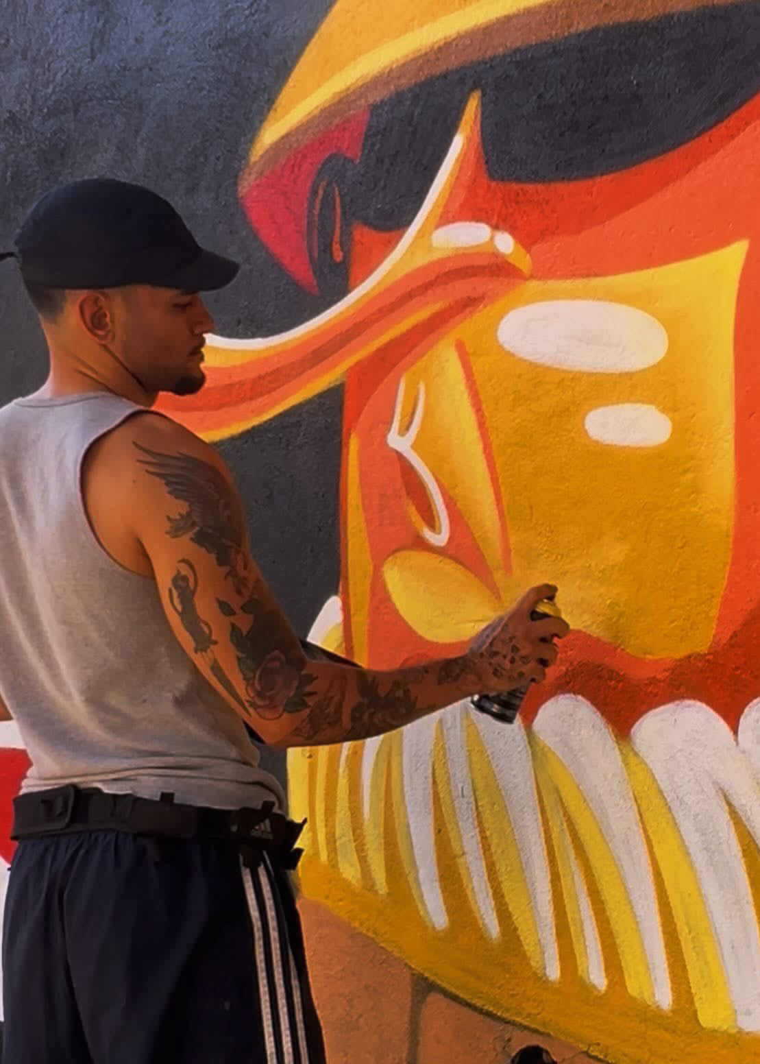 A man wearing a black baseball cap, gray tank top, and black athletic shorts with white stripes is spray painting a large colorful mural featuring a stylized face in yellow, orange, red, and white.