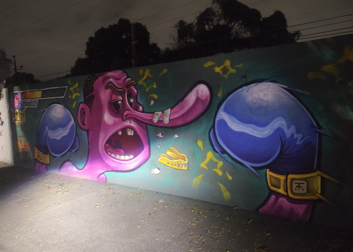 A large colorful graffiti mural painted on a wall at night, featuring a cartoon face with a long tongue, blue boxing gloves, and yellow star-like accents, photographed from below in an outdoor alley.