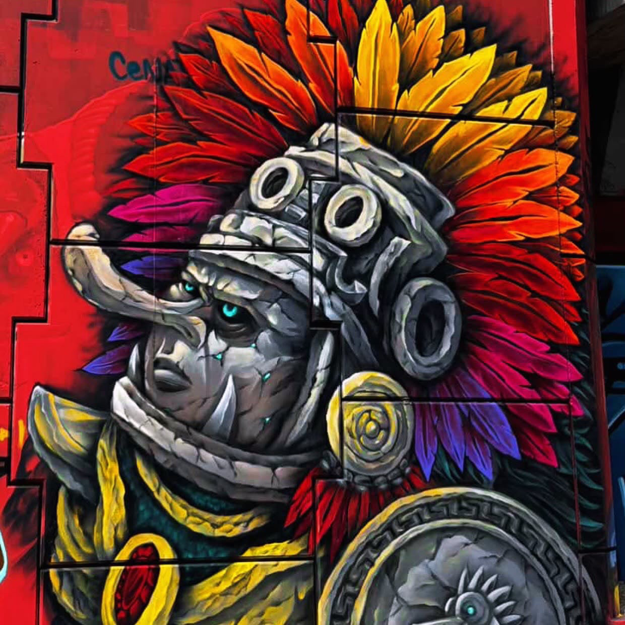 Colorful graffiti-style mural artwork showing a stylized human face in profile with a gray stone-like helmet and bright feathered headdress in red, orange, yellow, purple, and pink, set against a red background.