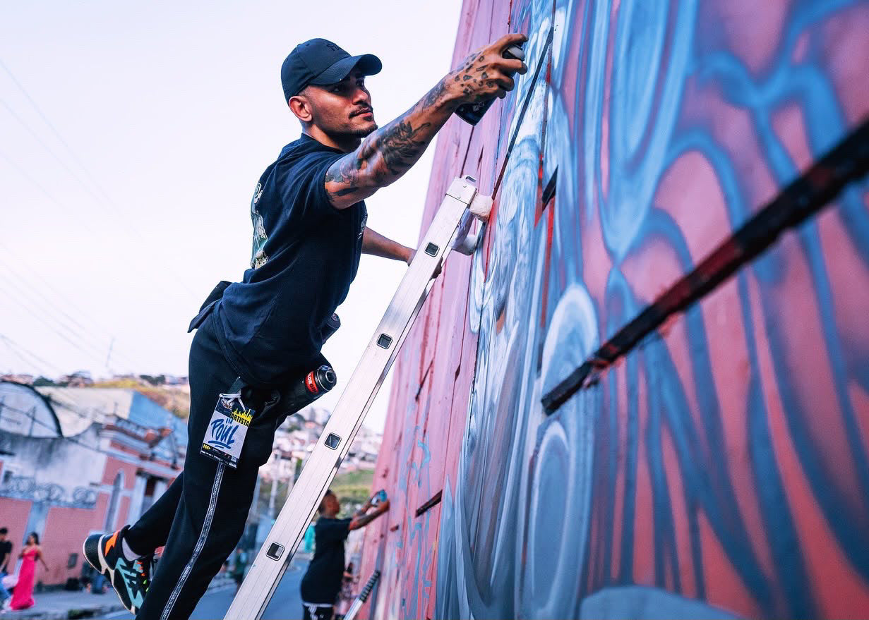A tattooed man in a black cap and black t-shirt climbs a metal ladder to paint a large mural on a red and blue wall during the day.