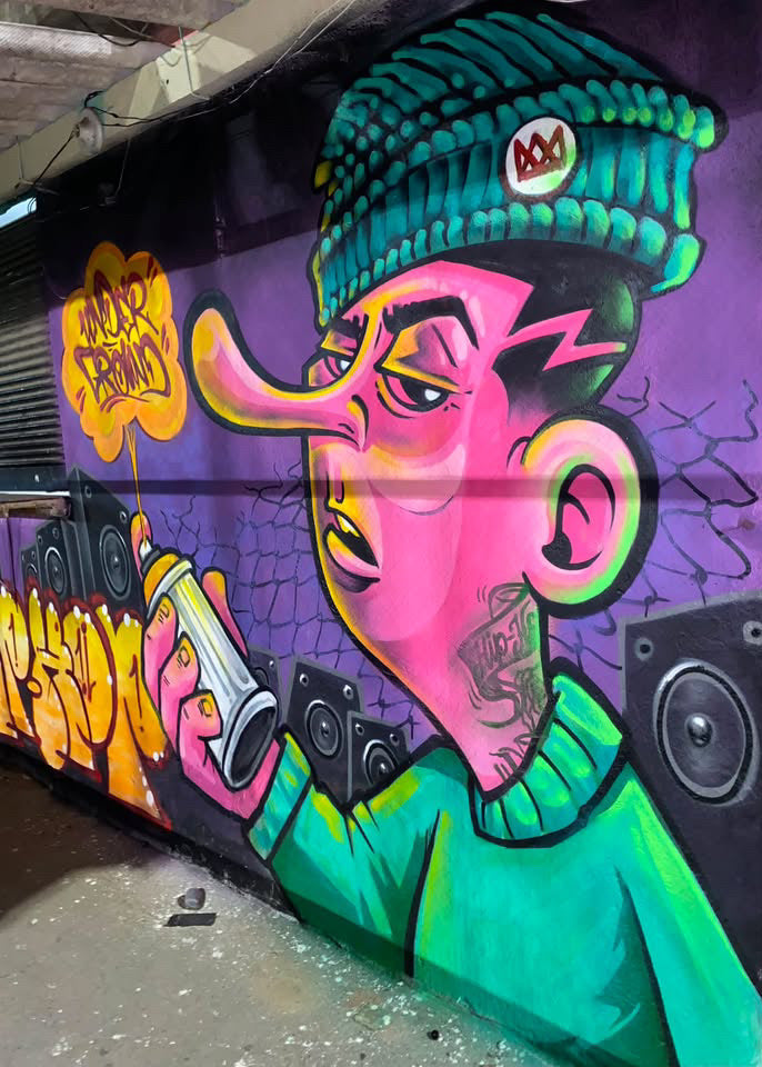 A colorful graffiti mural painted on a wall, featuring a stylized cartoon character with a pink face, green hat and sweater, holding a spray can, set against a purple background with yellow graffiti text.