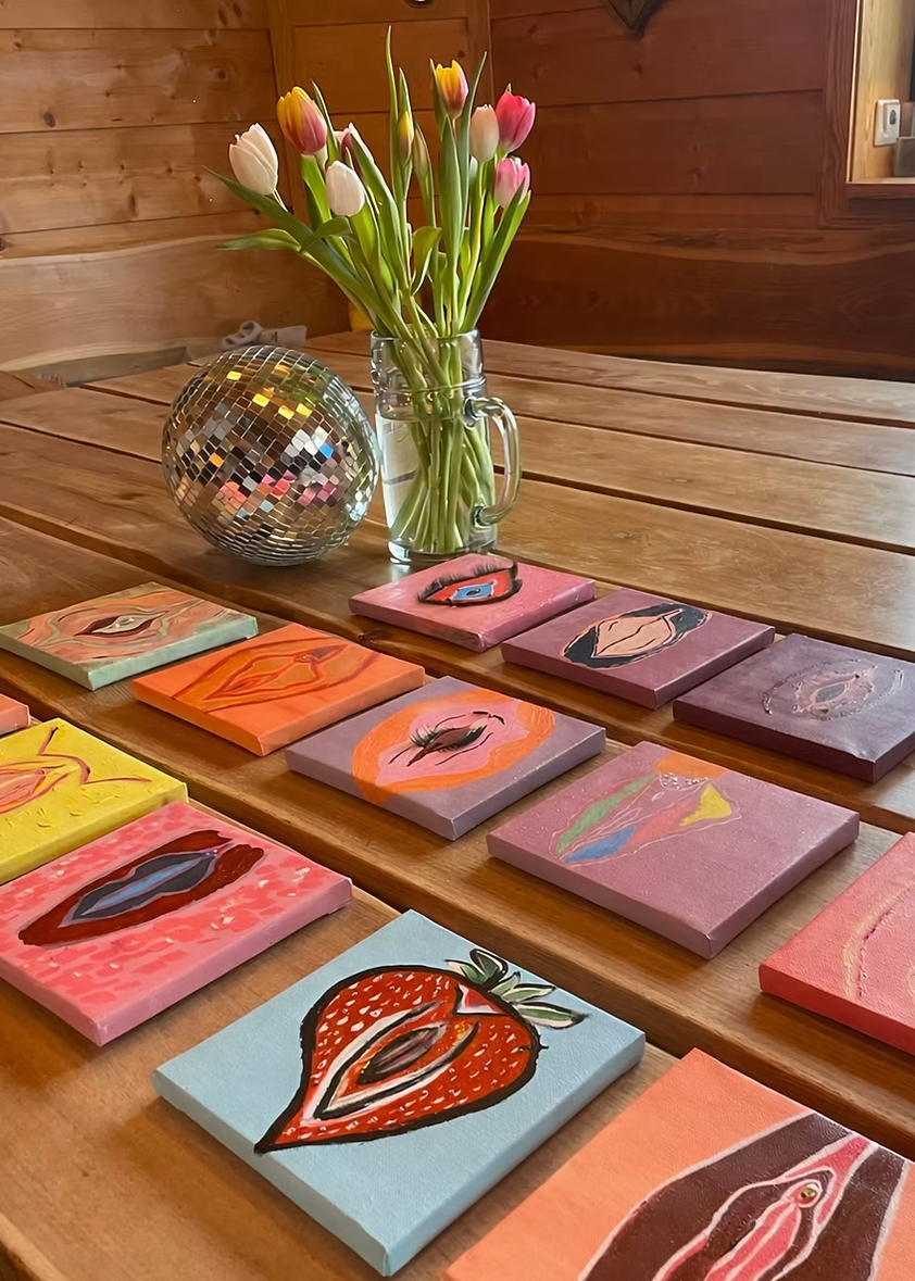 A collection of colorful hand-painted square canvas art pieces depicting stylized lips and eyes, arranged on a wooden table next to a glass jar of tulips and a disco ball in a rustic wood-paneled room.