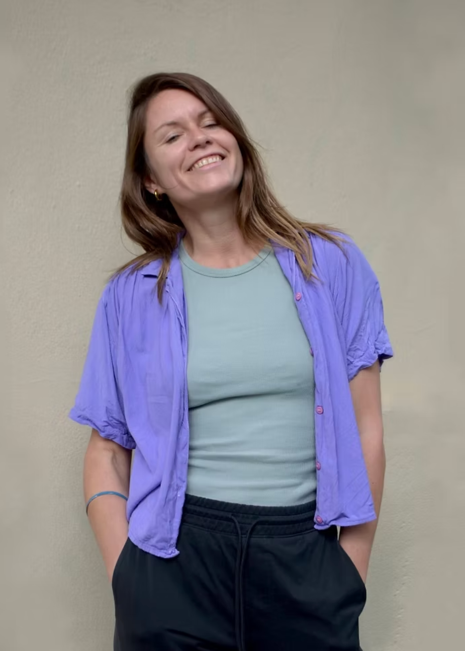 A woman wearing a light purple short-sleeve button-up shirt over a sage green crewneck top and black sweatpants, standing against a plain beige wall.