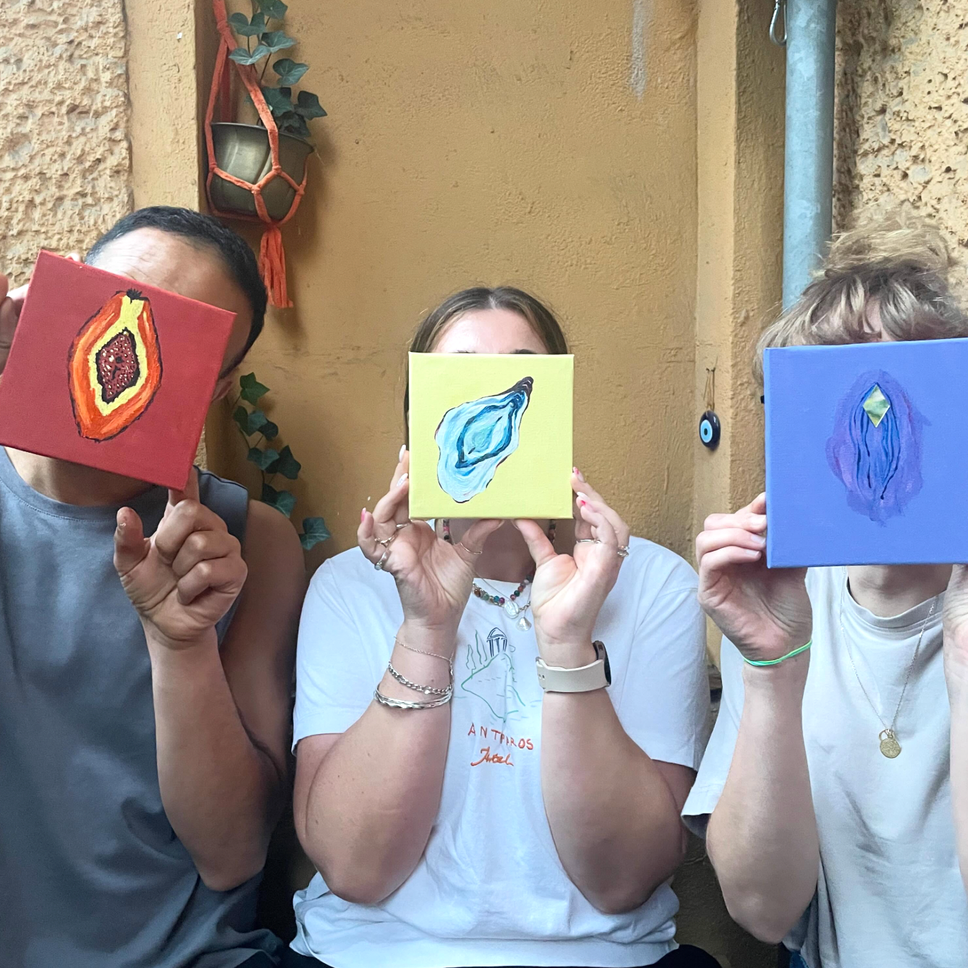 Three people holding up small square paintings with colorful abstract designs, set against a warm beige stucco wall background.