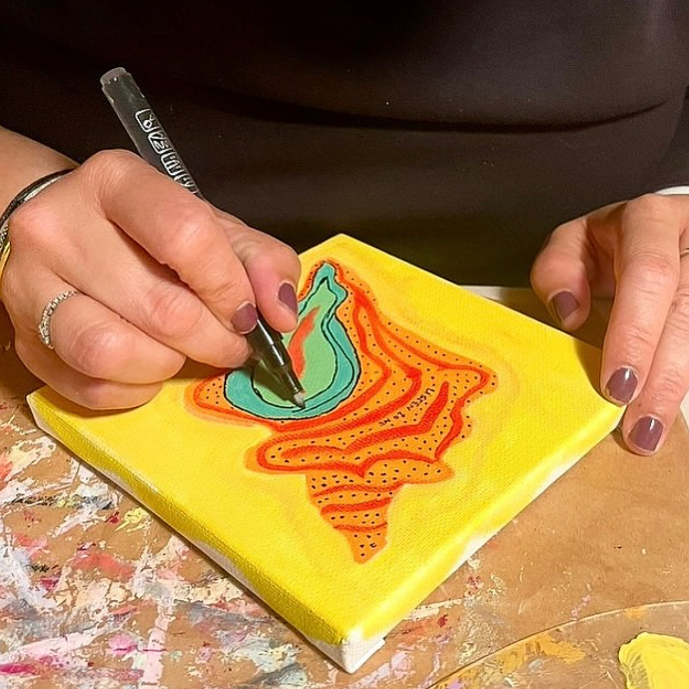 A close-up of a person's hands painting on a small square yellow canvas. The hand holding a fine-tip paintbrush applies teal and orange paint to an abstract design.