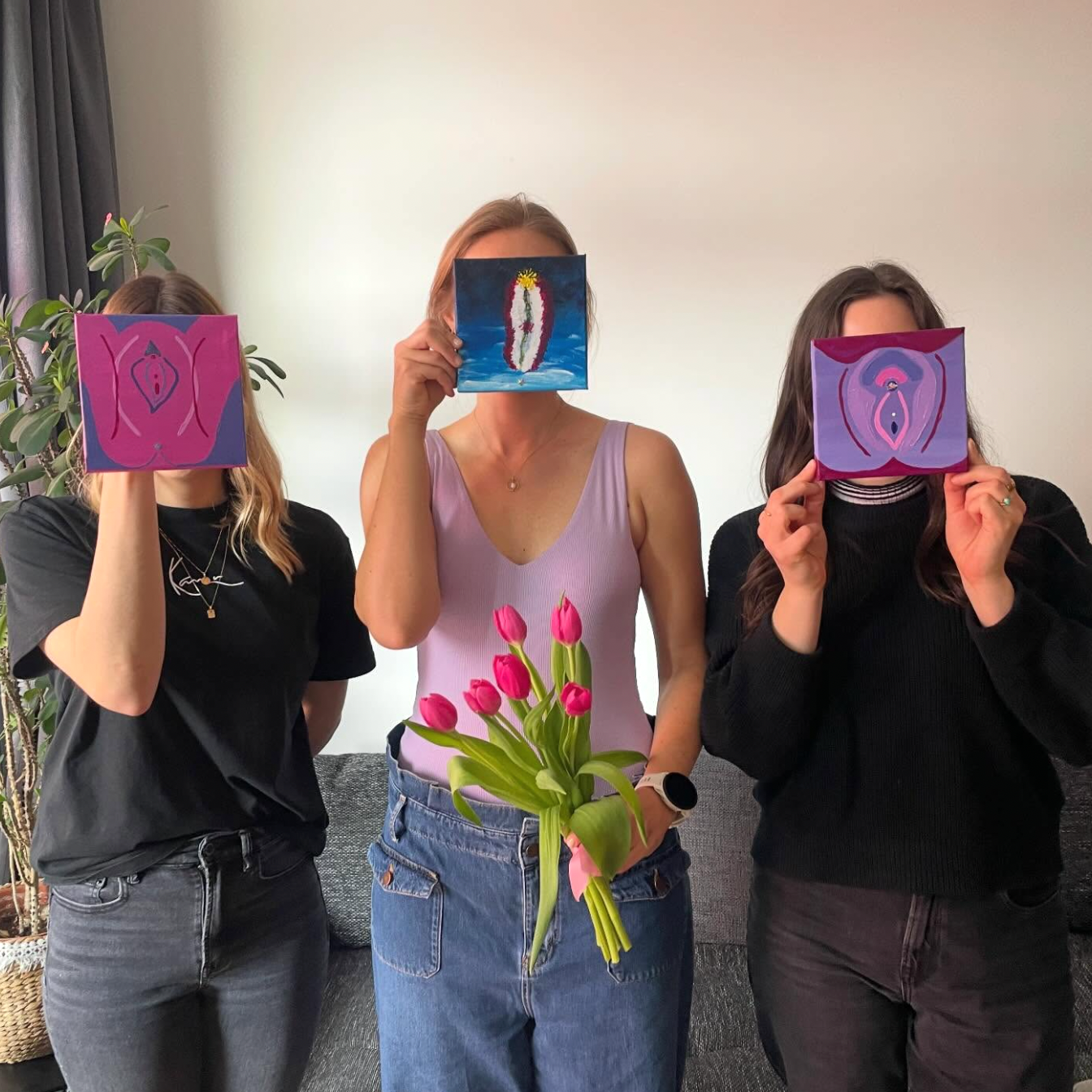 Three women standing indoors in front of a plain wall, each holding a small square artwork featuring a stylized pink and purple abstract face. One woman in the center also holds a bouquet of pink tulips.