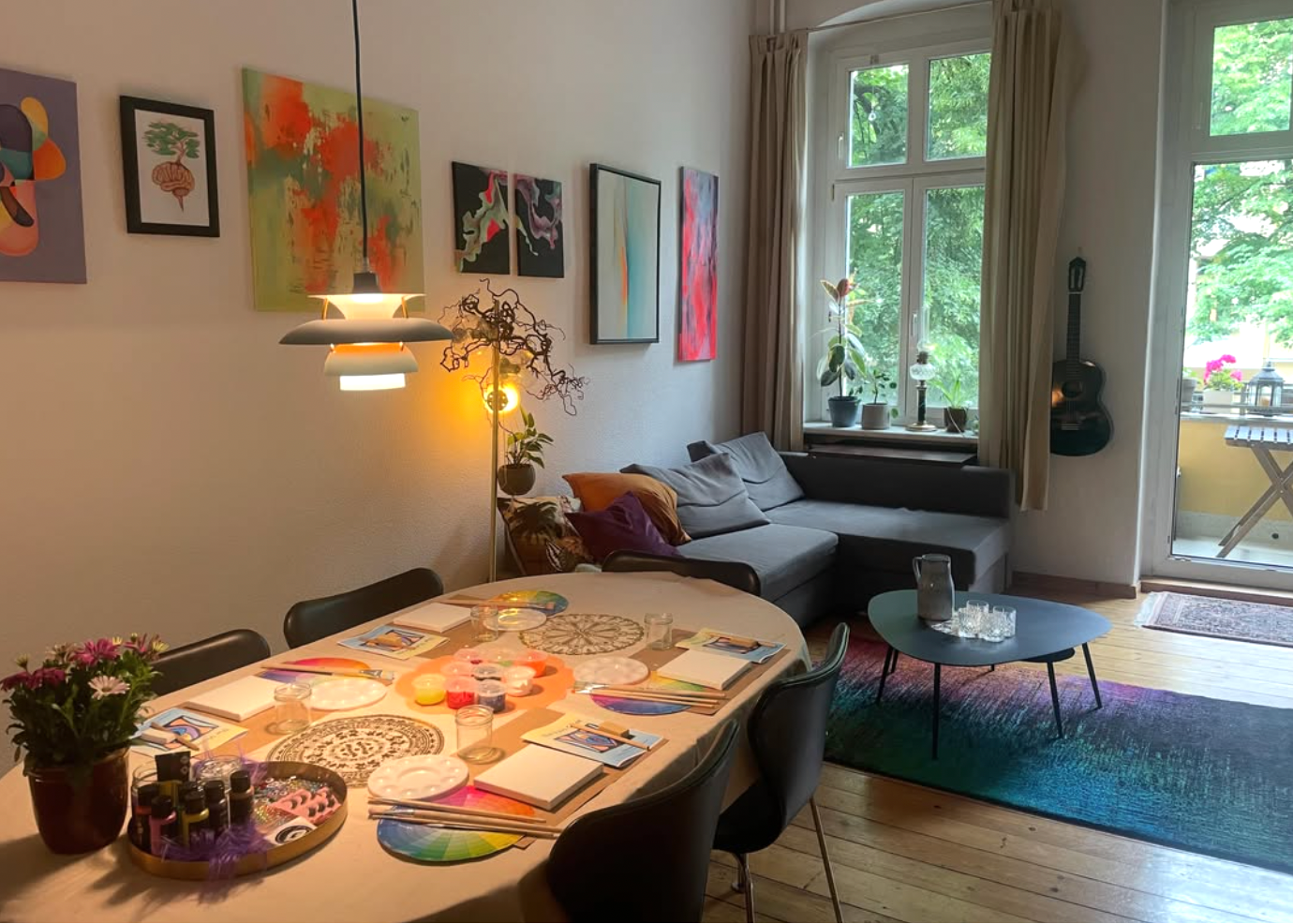 A bright, modern living room with a gray sectional sofa, a round black coffee table, a colorful gradient rug, and a dining table with chairs. The room features multiple colorful abstract paintings on the wall, a hanging pendant light, and large windows looking out onto green trees.
