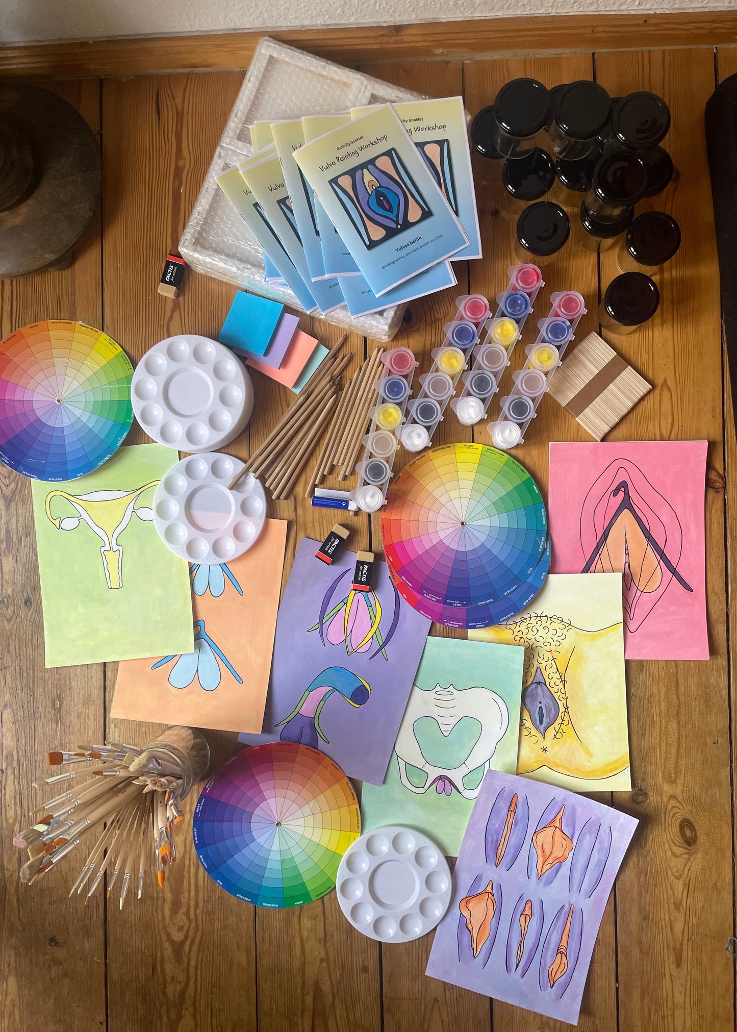An overhead flat-lay of a colorful art and painting supplies set arranged on a wooden floor. The collection includes multiple circular color wheel cards, white plastic paint palettes, small tubes of acrylic paints, wooden sticks, paper, and several finished abstract art prints in pink, yellow, blue, and purple.