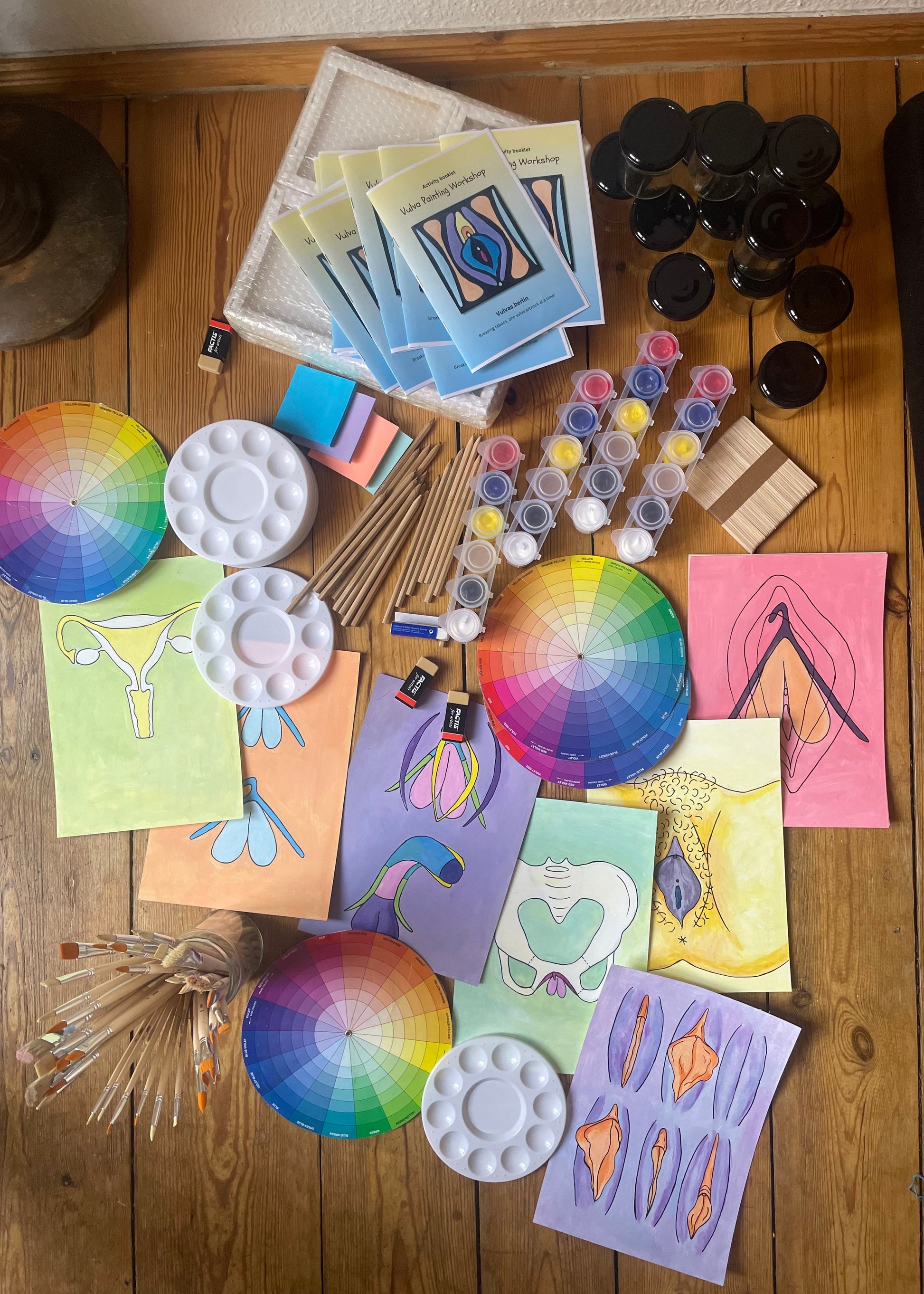 An overhead flat-lay of a colorful art and painting supplies set arranged on a wooden floor. The collection includes multiple circular color wheel cards, white plastic paint palettes, small tubes of acrylic paints, wooden sticks, paper, and several finished abstract art prints in pink, yellow, blue, and purple.