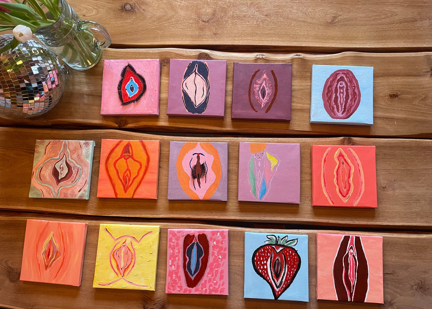 A set of 15 small square canvas paintings arranged in three rows on a wooden table. Each canvas depicts a stylized female vulva in various colors and artistic styles.