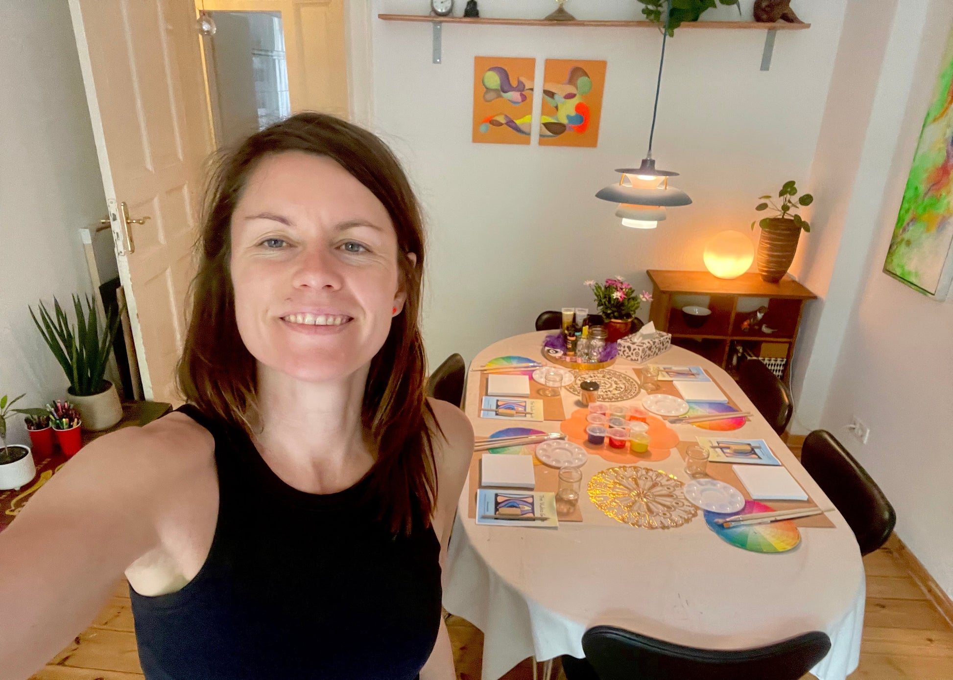 A woman taking a selfie in a brightly lit home interior, with a colorful art and craft table set up in the background. The table features various colorful materials, papers, and small items, and the room has warm lighting from a hanging lamp and a glowing lamp on a side table.