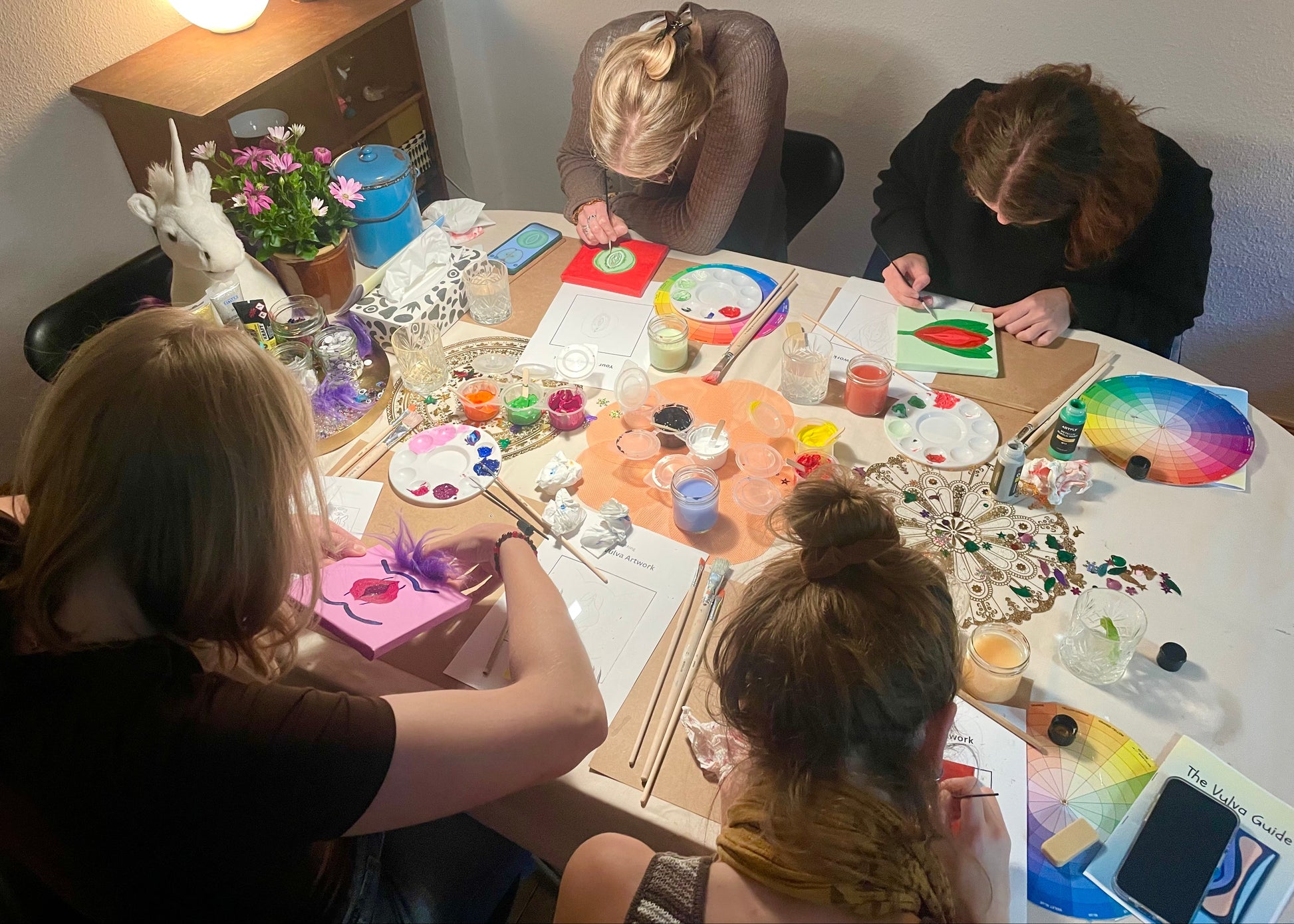 Four people sitting around a round table, painting and crafting with various art supplies, paint palettes, brushes, and colorful materials in a warmly lit indoor setting.