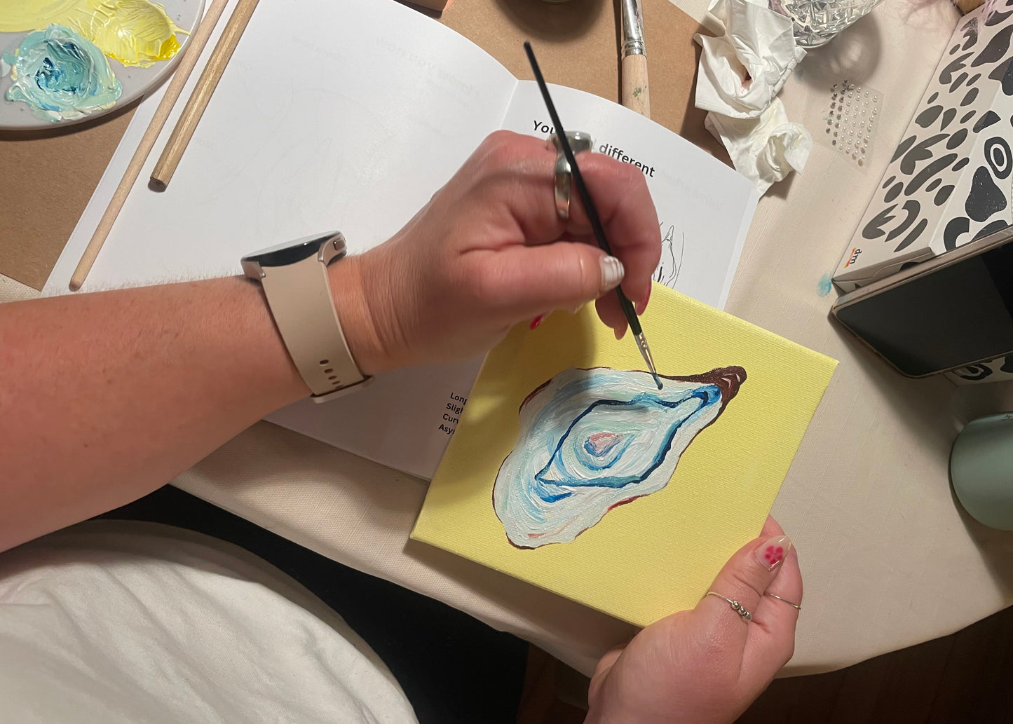 A person's hands holding a small yellow canvas painting of an oyster shell, using a fine paintbrush to apply blue and brown paint, set in a creative workspace with art supplies.